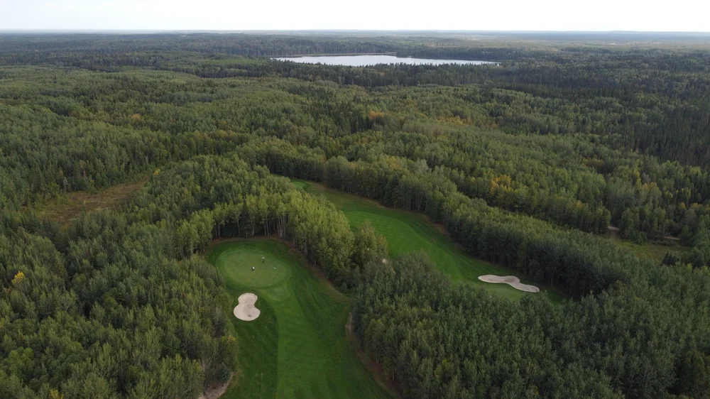 Aerial view of a lush green golf course surrounded by boreal forest with a lake beyond