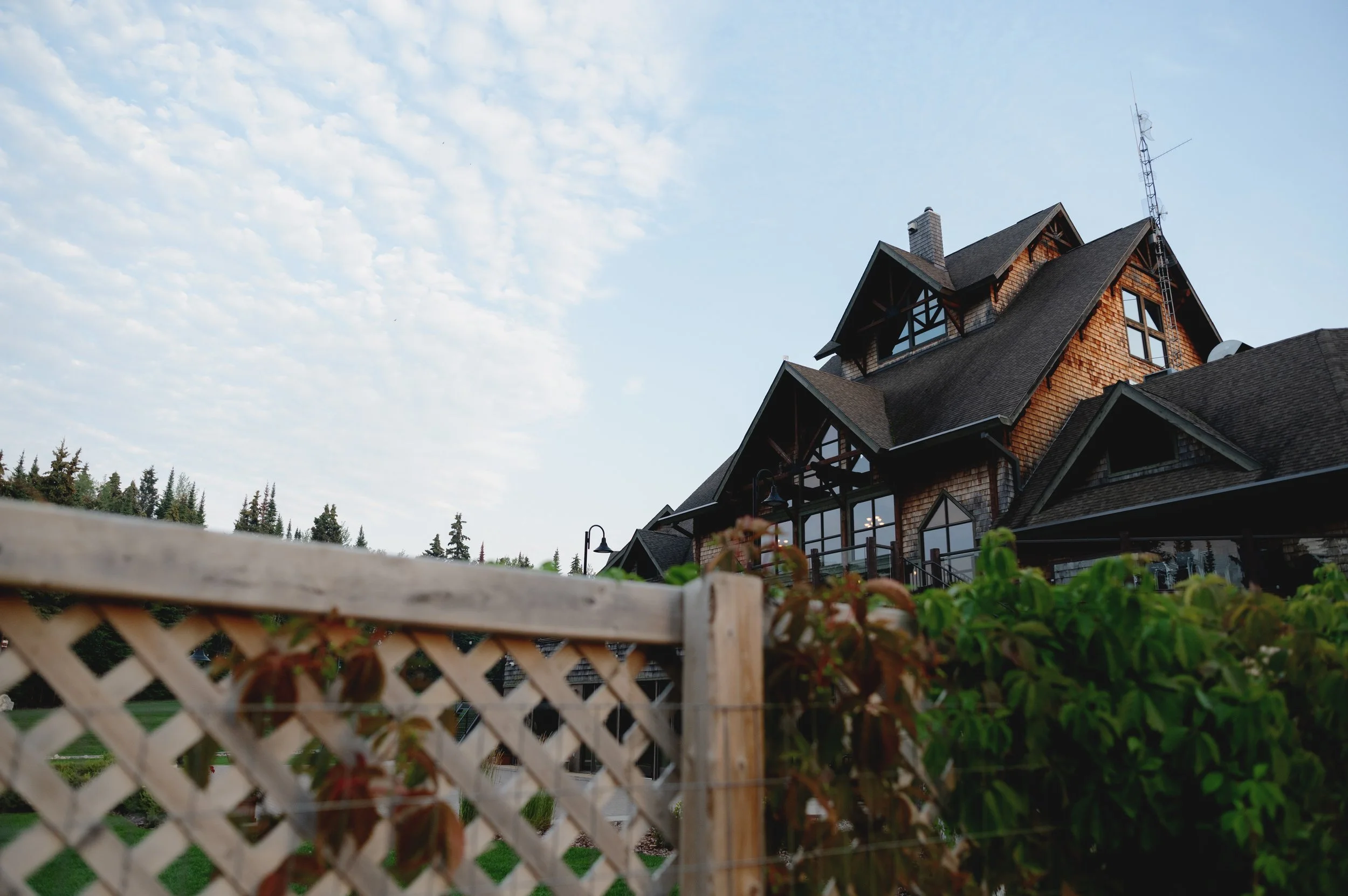 A large house with a steep gabled roof, wooden shingles, and multiple windows, set against a blue sky with scattered clouds. A wooden fence is visible in the foreground.