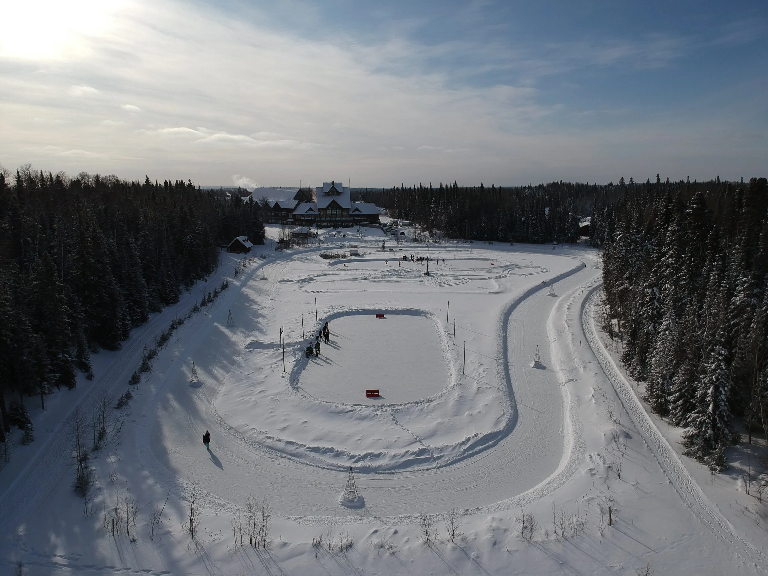 A snowy landscape with a building in the distance surrounded by trees and a cloudy sky. There are people and sleds on the snow, and ski tracks and paths are visible.