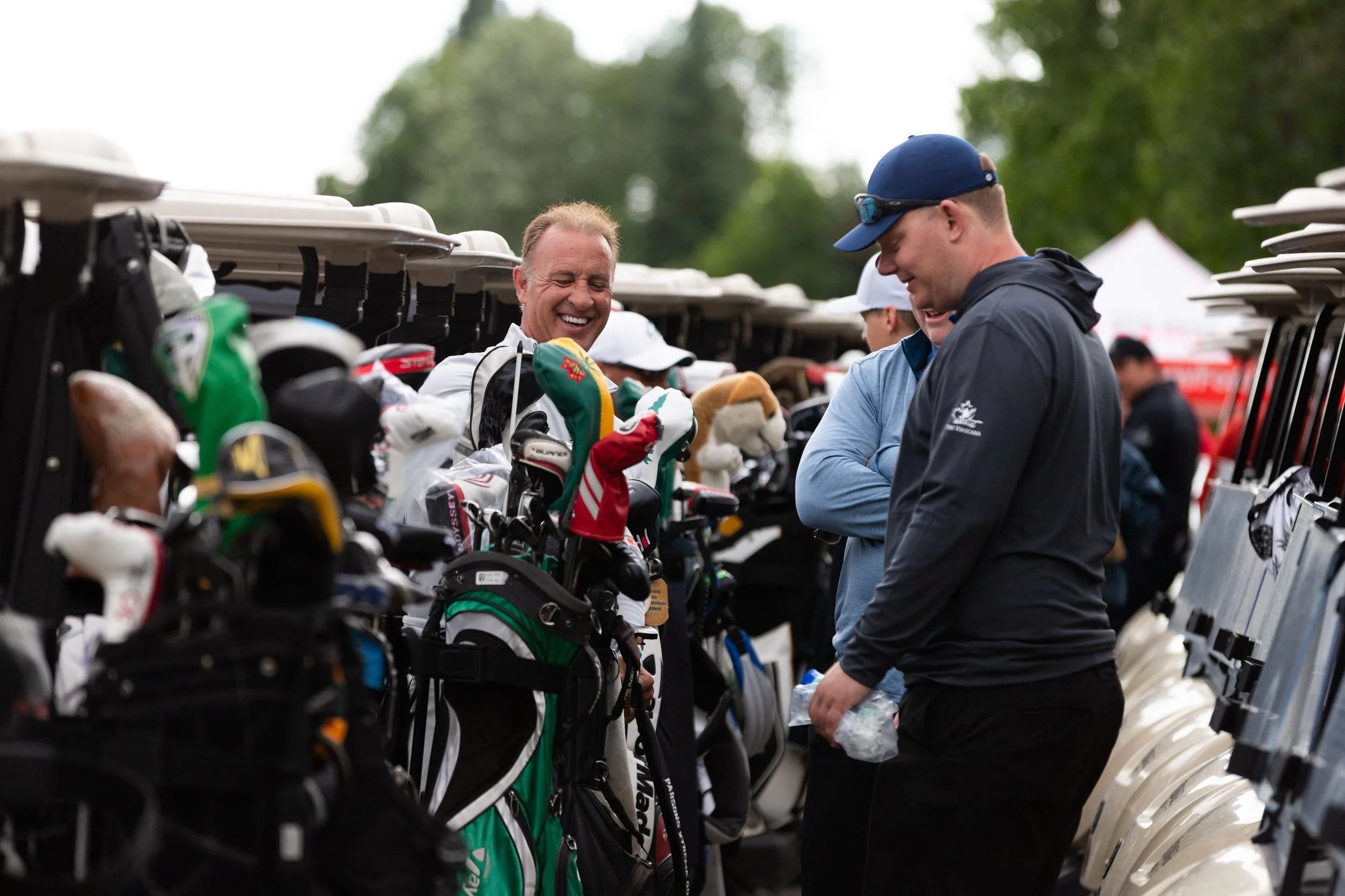 Two men are smiling and talking at a golf cart rack filled with golf clubs and equipment, outdoors on a cloudy day.