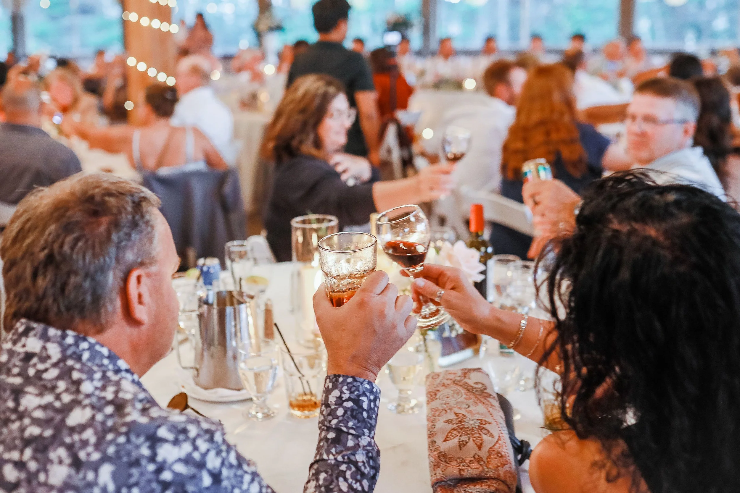A group of people gathered around a table at a social gathering, holding drinks and engaging in conversation, with some toasting each other.