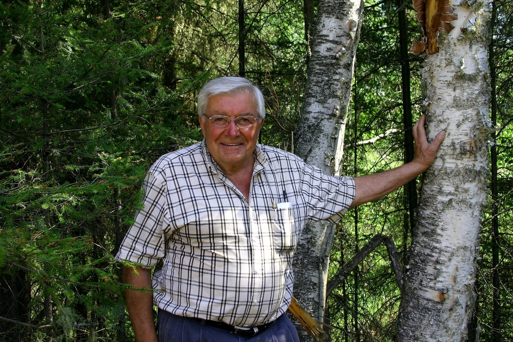 An elderly man in a checkered shirt smiling and standing outdoors in a forest, with his hand resting on a birch tree.