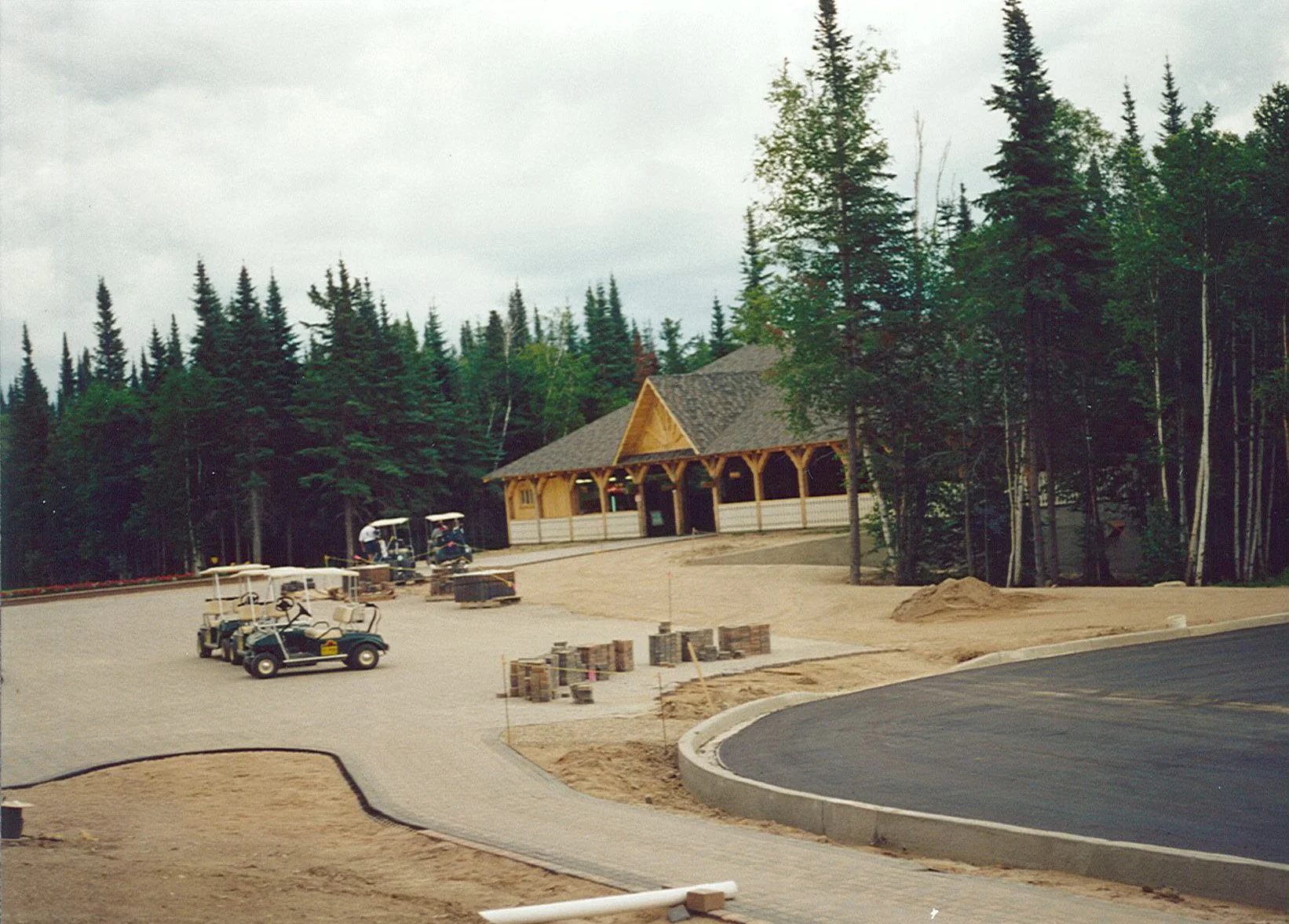 A parking lot under construction with golf carts, stacks of bricks, a building with wooden accents, and trees in the background.
