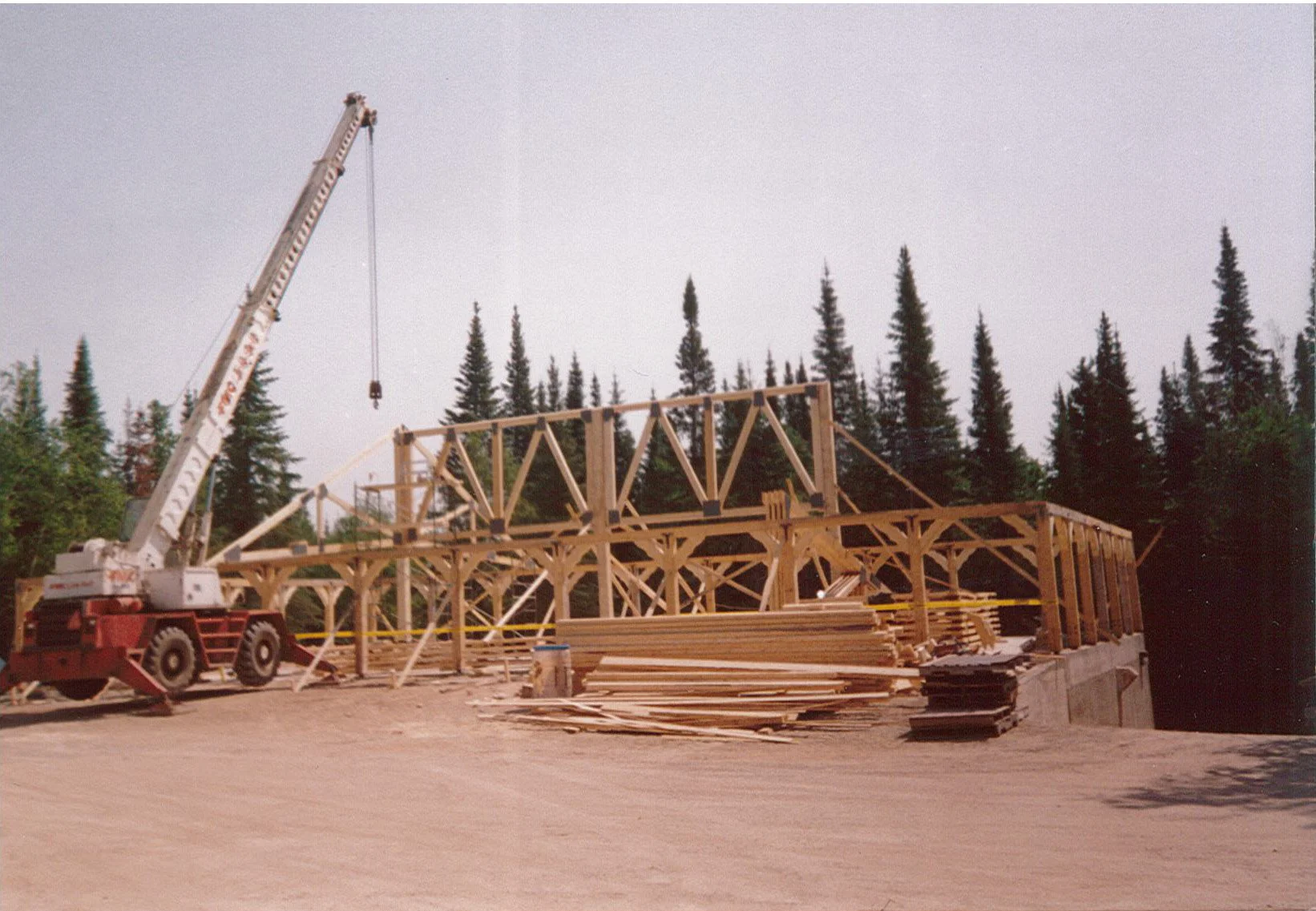 Construction site featuring a wooden framework for a building, a crane on the left, and stacks of lumber, with trees in the background.