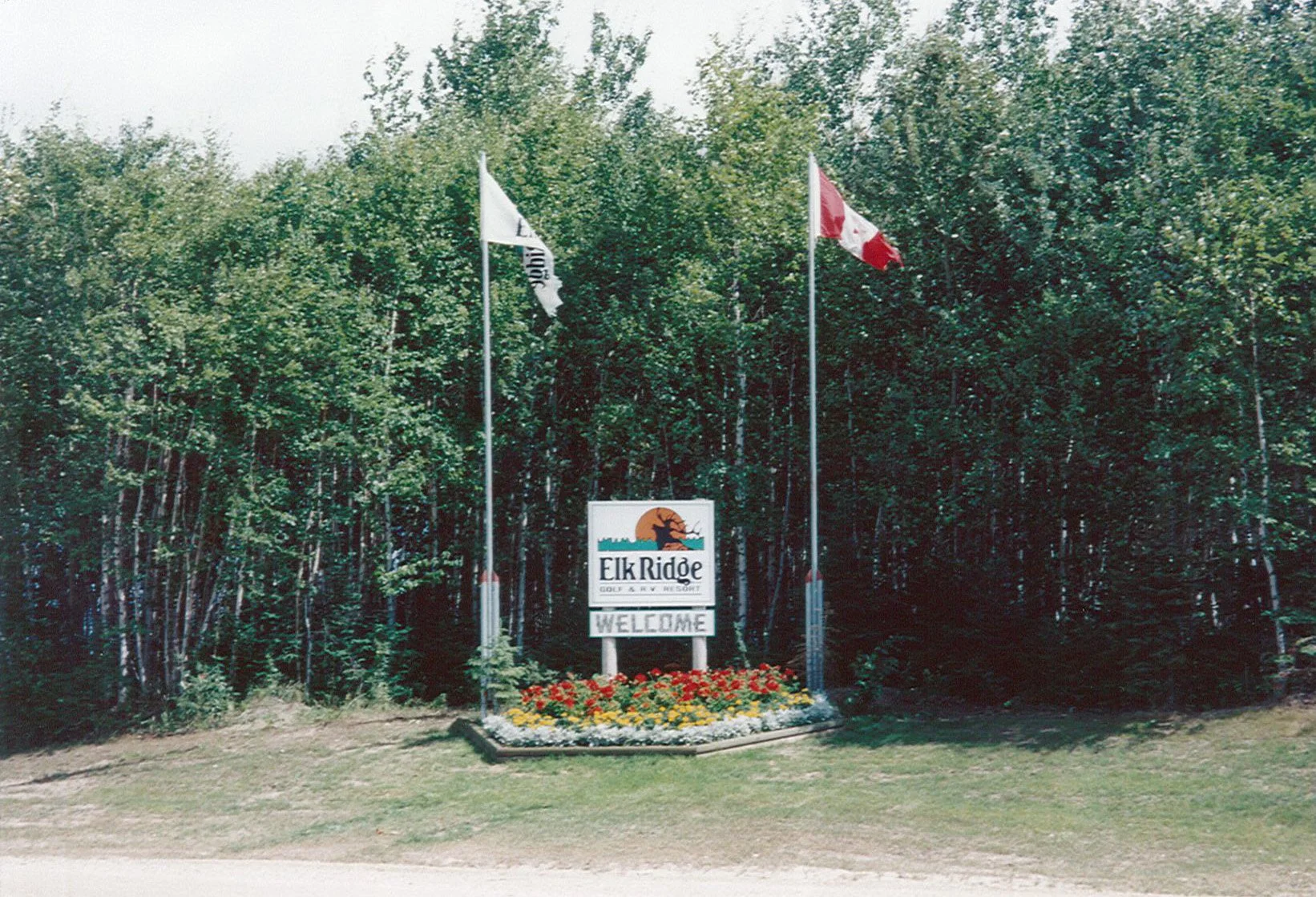 Entrance sign for Elk Ridge, a golf and ski resort, with a landscaped flower bed in front, and two flags flying, one of which is the Canadian flag. Dense trees surround the area.