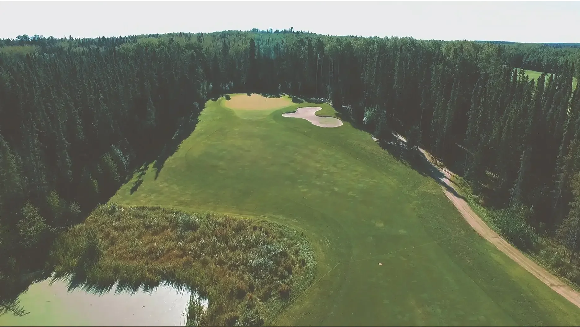 Aerial view of a golf course fairway with a sand trap, surrounded by dense forest, and a gravel pathway on the right.