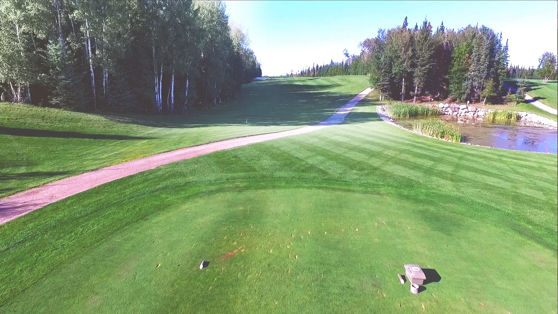 A golf course with a grassy tee box, a fairway, trees left and right, a small pond on the right side, and a clear blue sky.