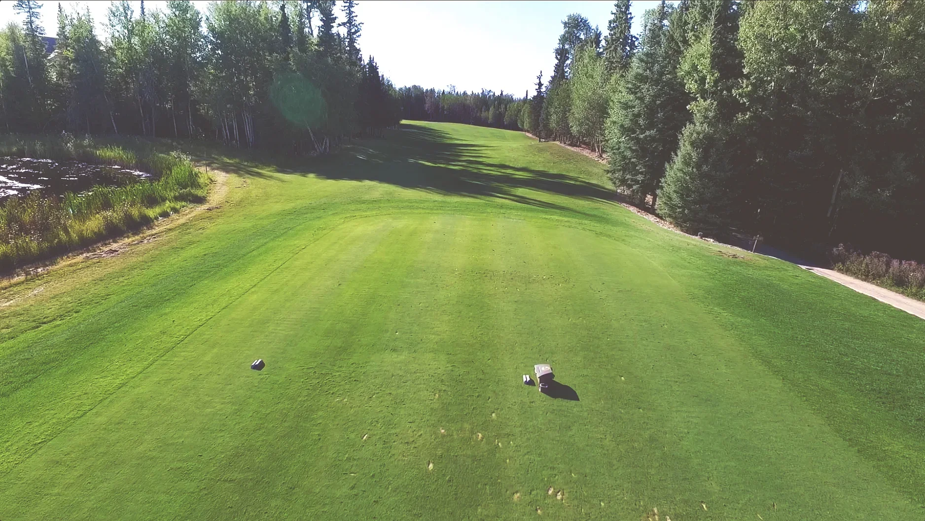View from a golf tee box looking down a fairway surrounded by trees, with golf clubs on the ground.