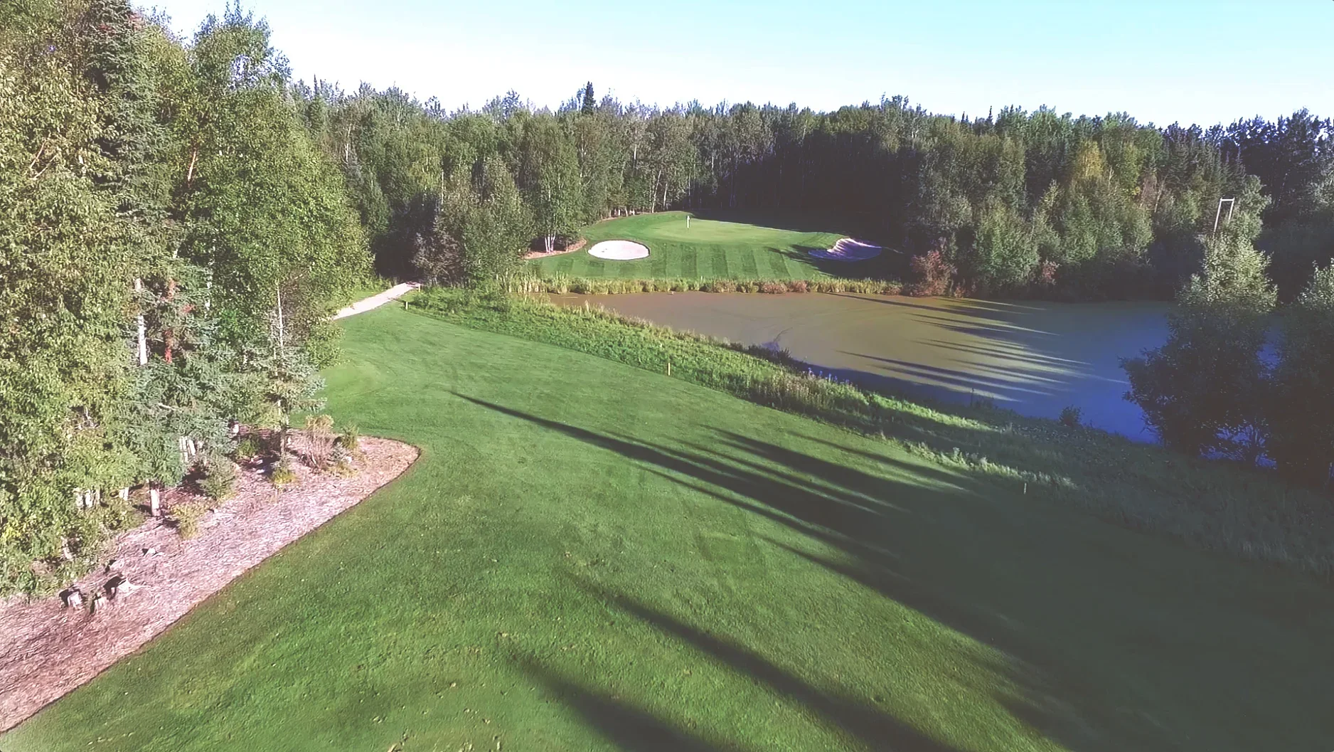 A golf course with a green fairway, sand bunkers, a small pond, and trees surrounding the area under a clear blue sky.