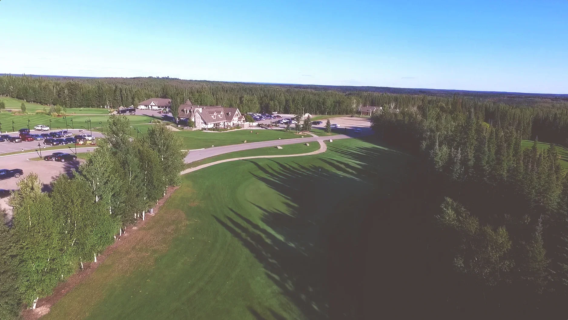 Aerial view of a golf course with neatly mowed green grass, trees lining the edges, a small building with a pitched roof, parking lots filled with cars, pathways, and a surrounding forest under a clear blue sky.