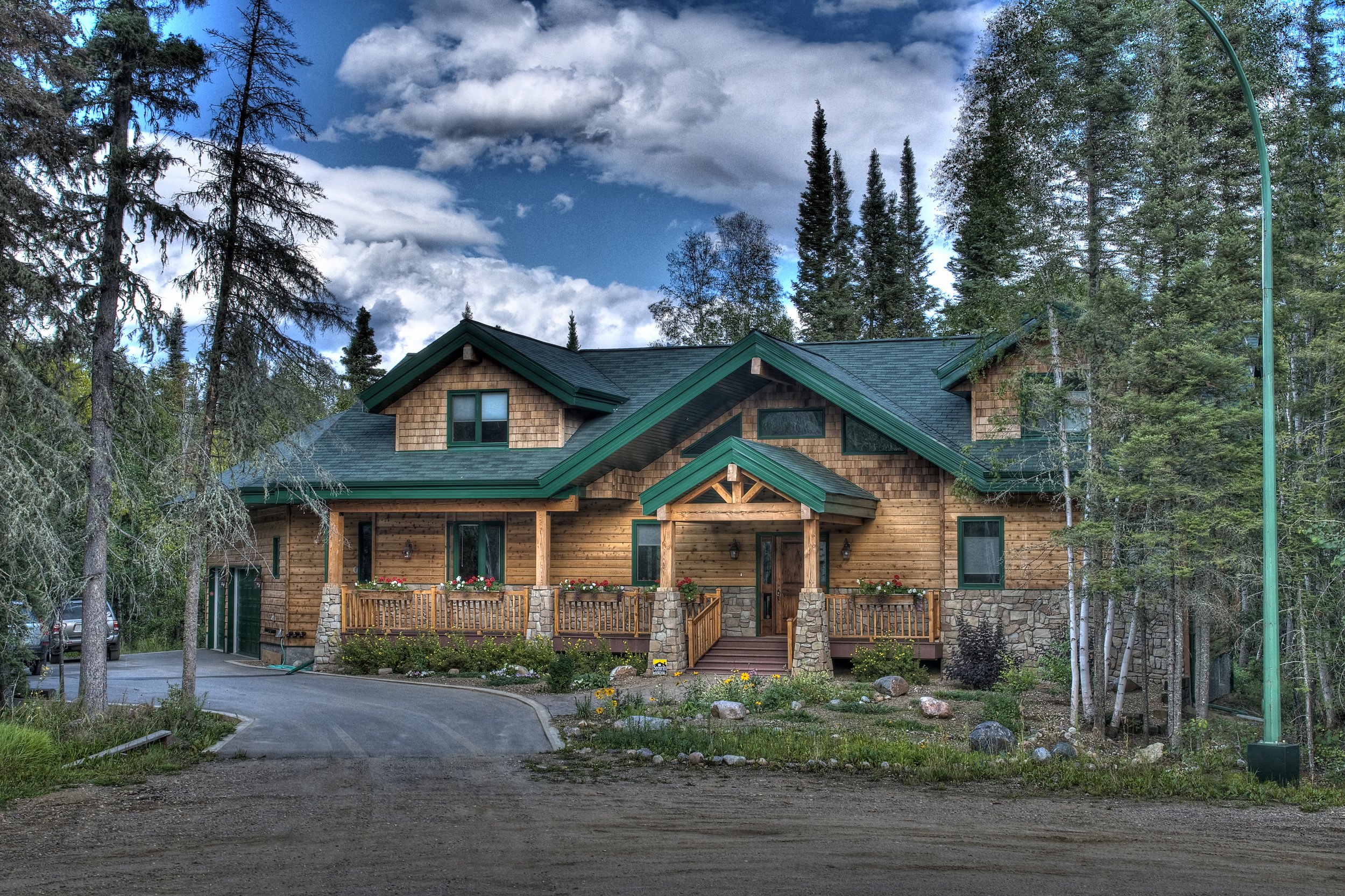 A wooden house with a green roof, surrounded by tall trees and a partly cloudy sky.