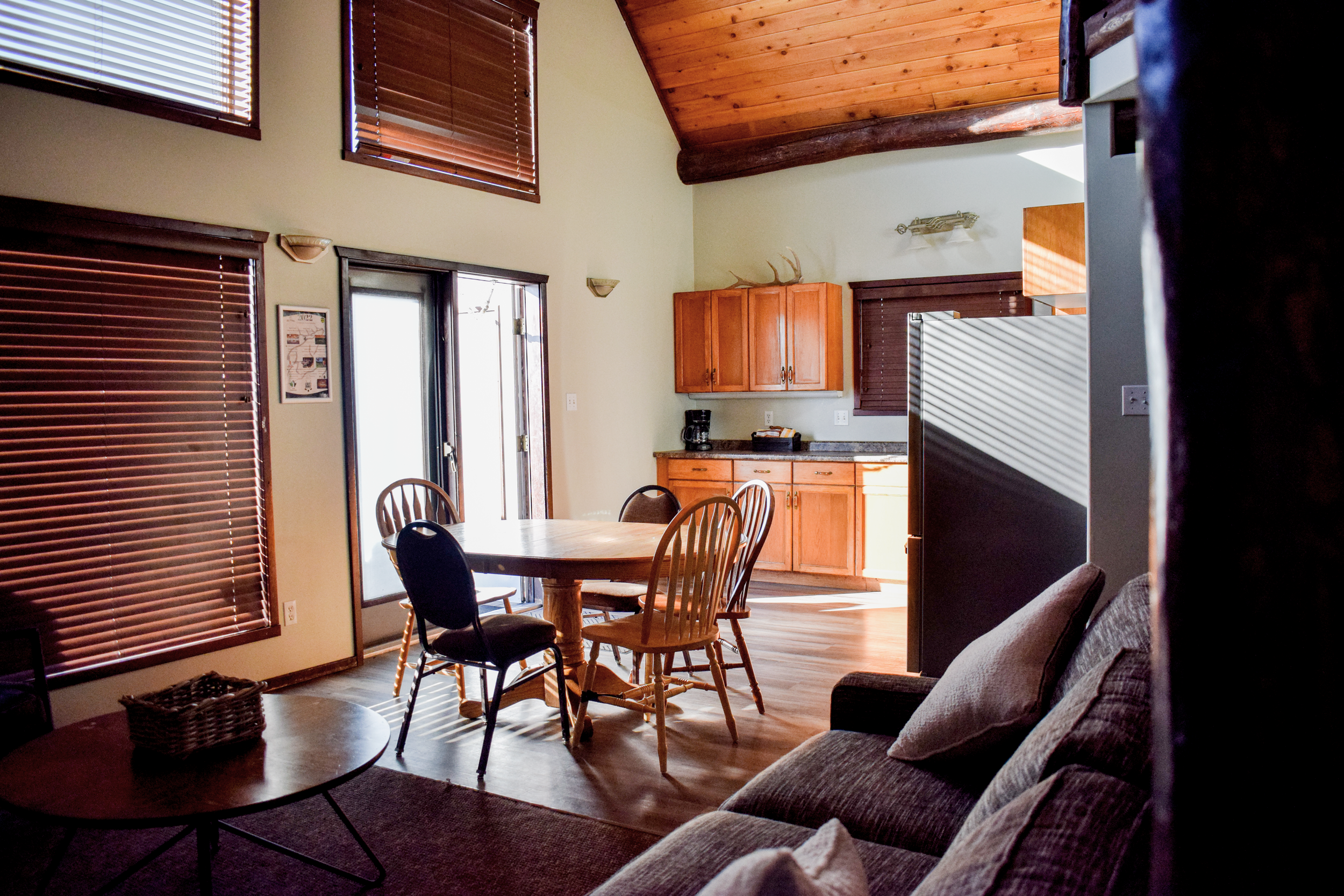 Interior view of a dining and living area with wood accents, including high wooden ceilings, furniture, and window blinds.