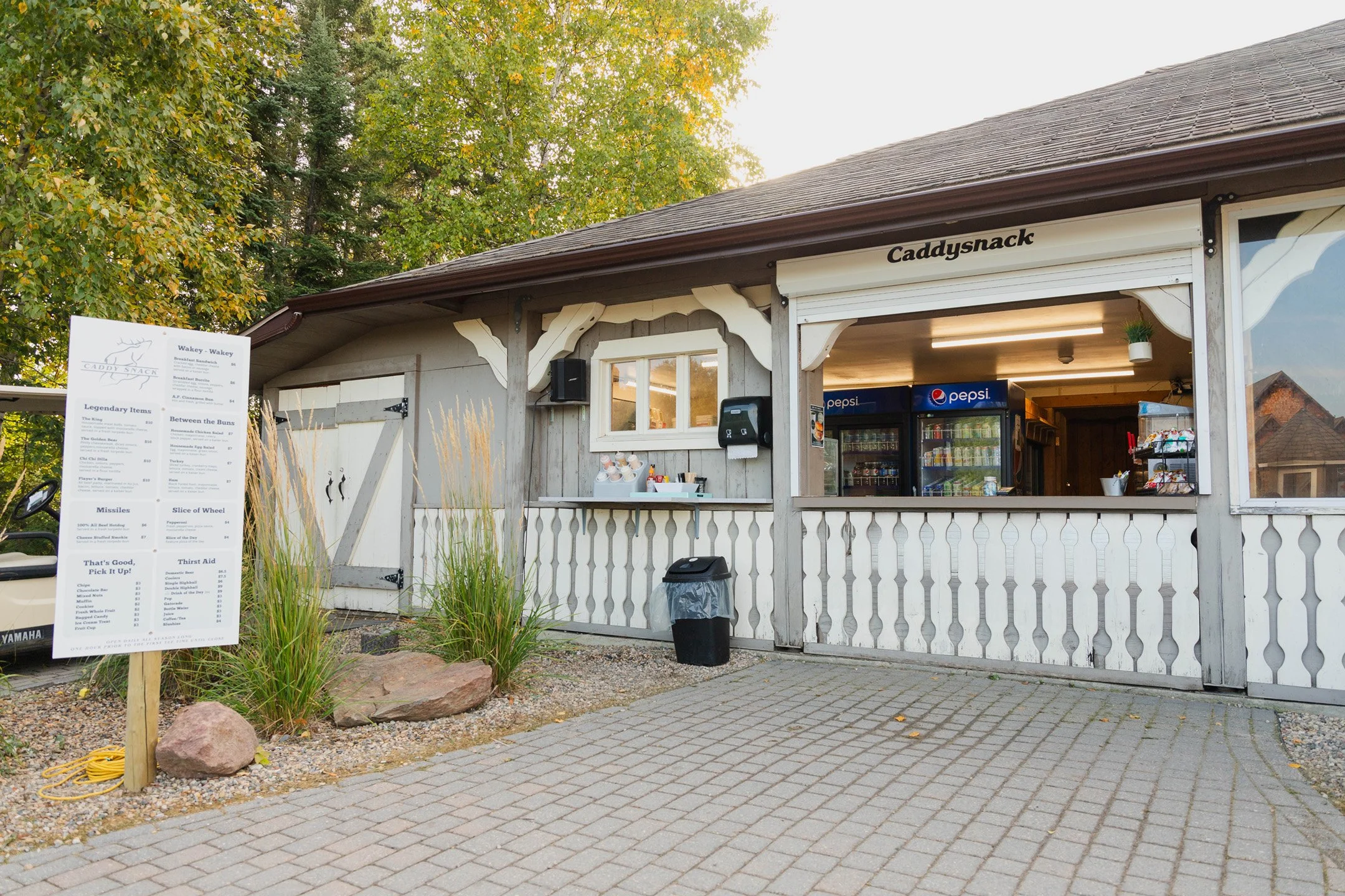 A small wooden snack stand with a menu board in front. The stand has a white railing, a small window, and a Pepsi refrigerator inside. There are trees with green and yellow leaves behind it, indicating fall season.