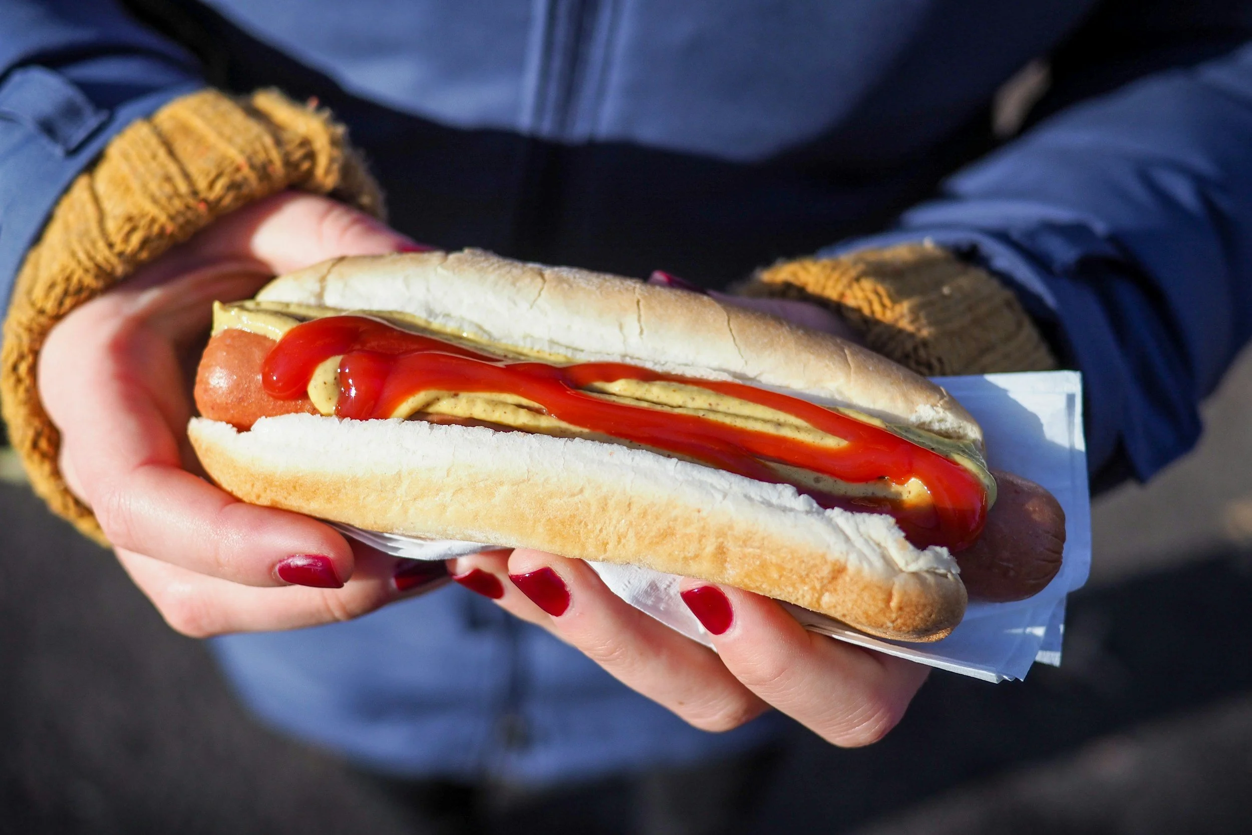 A person holding a hot dog in a bun with ketchup, mustard and sliced pickles.