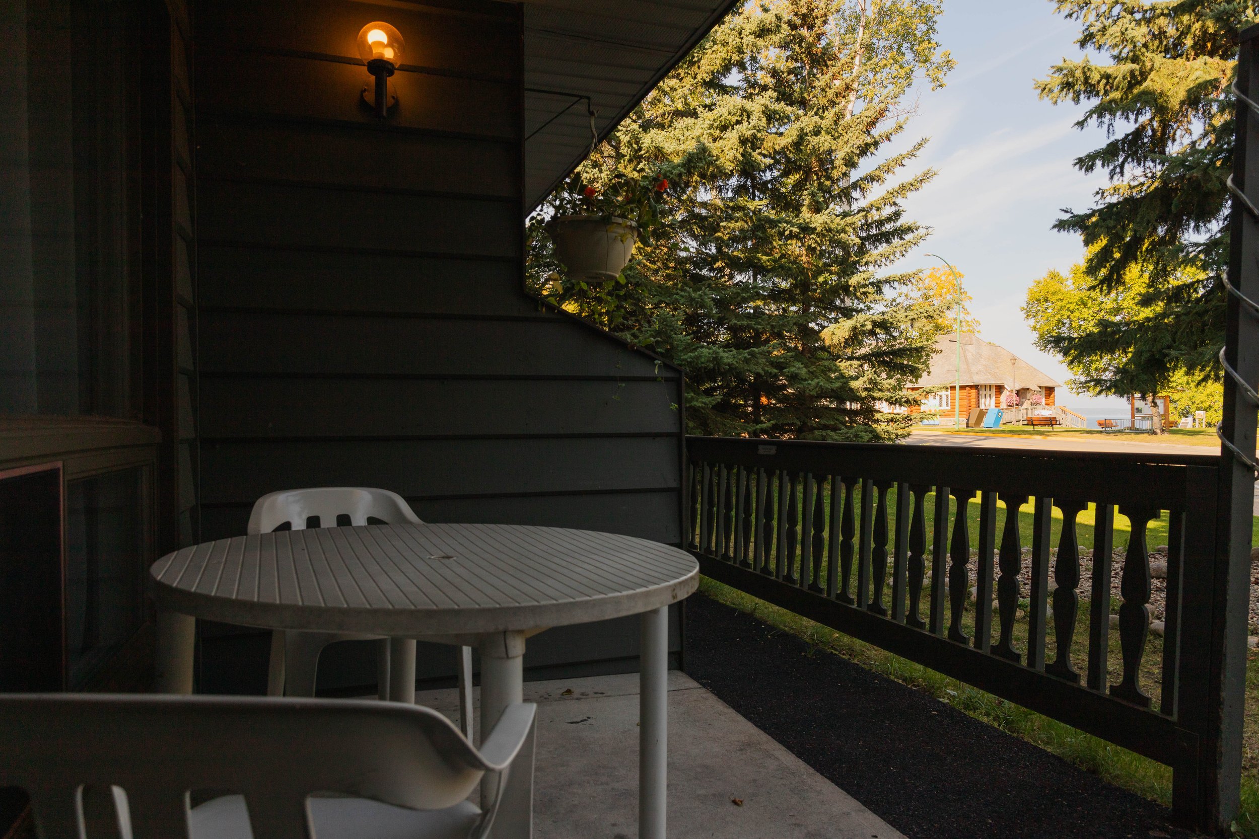 View from a porch showing a round plastic table and chairs, with a dark wood wall and a wall-mounted light fixture on the left, and a view of trees and a house in the background on a sunny day.