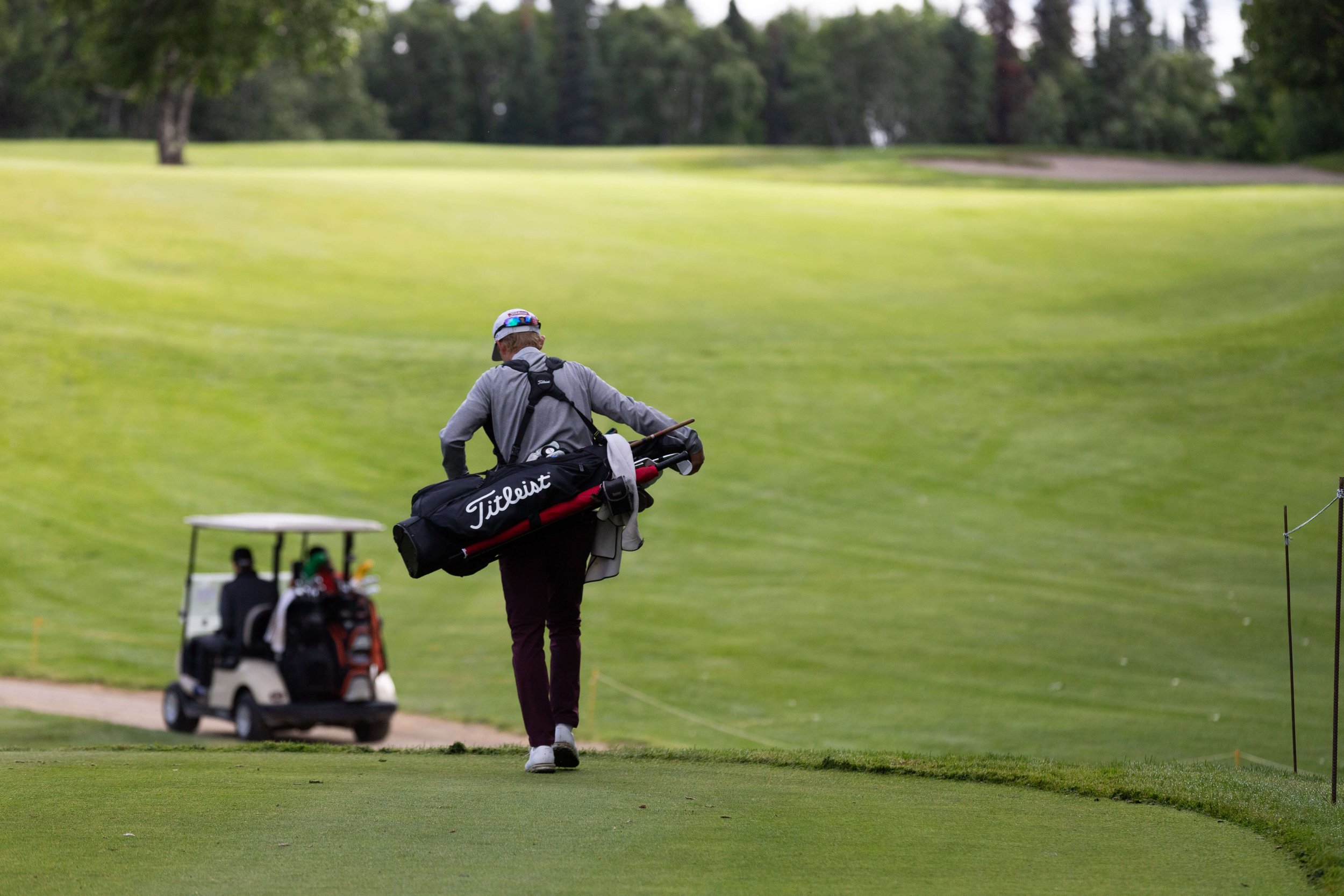 A golfer walking the fairway carrying a bag, with a power cart on the cart path behind.