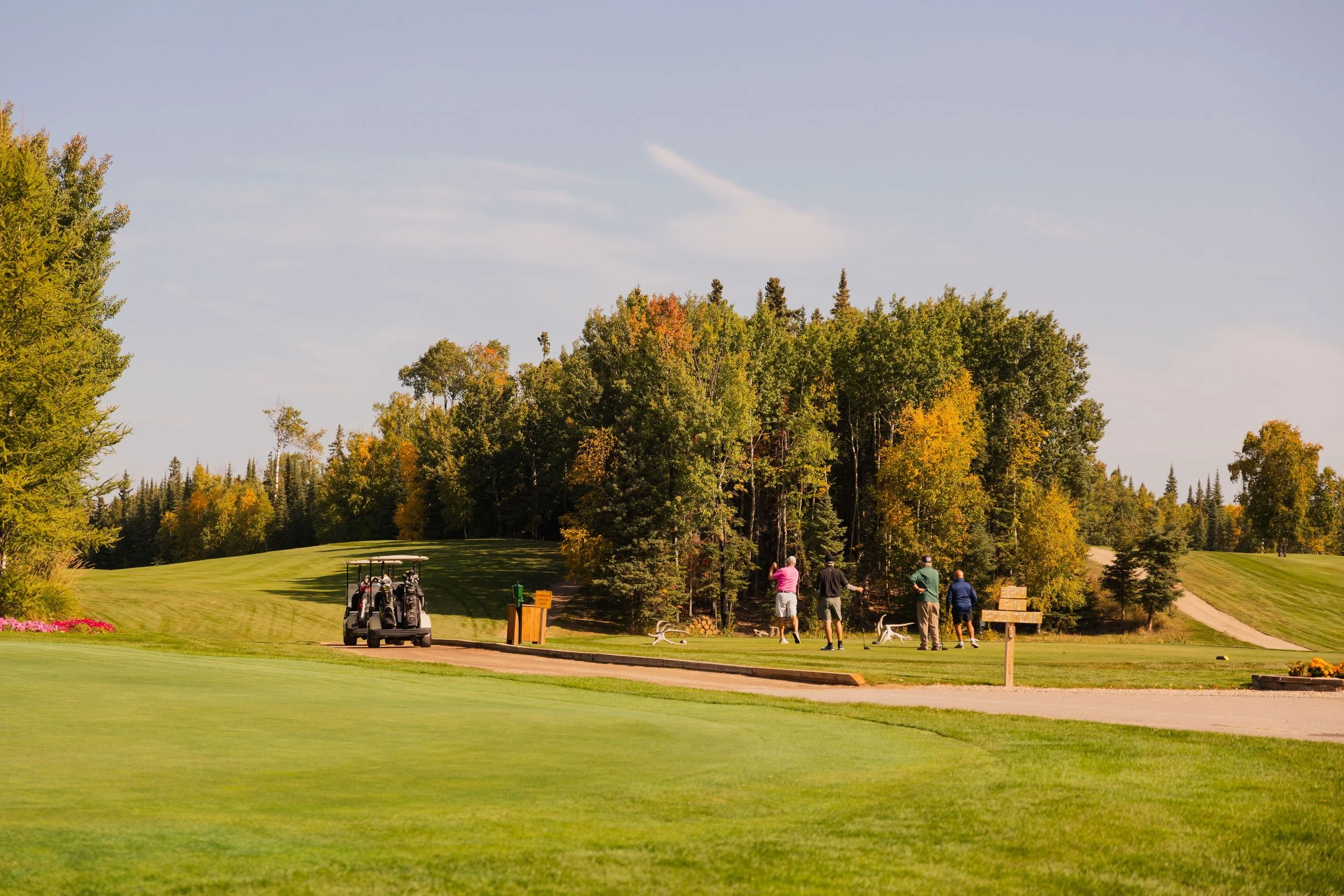 A golf course with four people playing, surrounded by trees with fall foliage, and golf carts parked nearby.