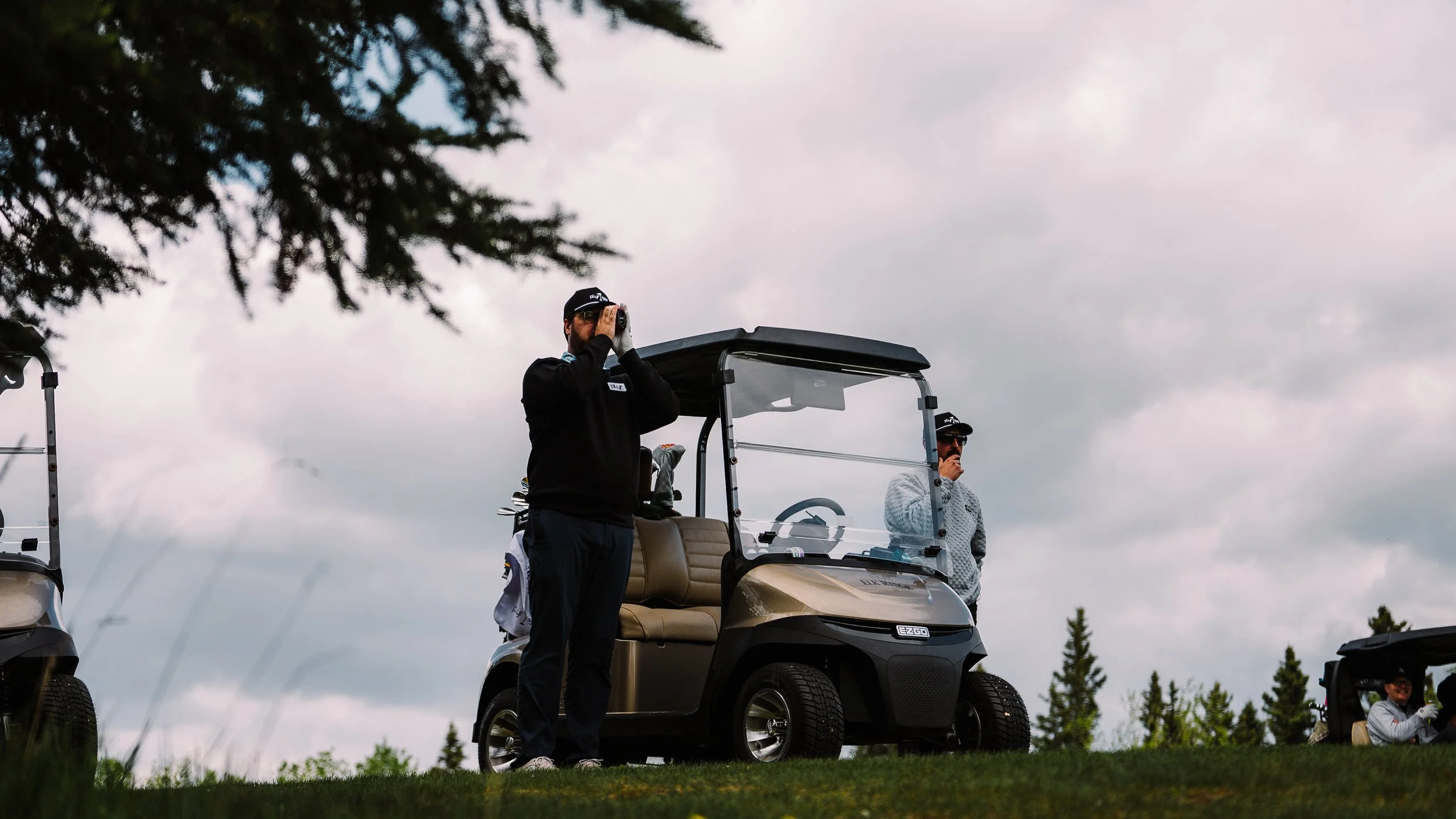 Two men standing near a golf cart on a cloudy day at a golf course, with trees in the background.
