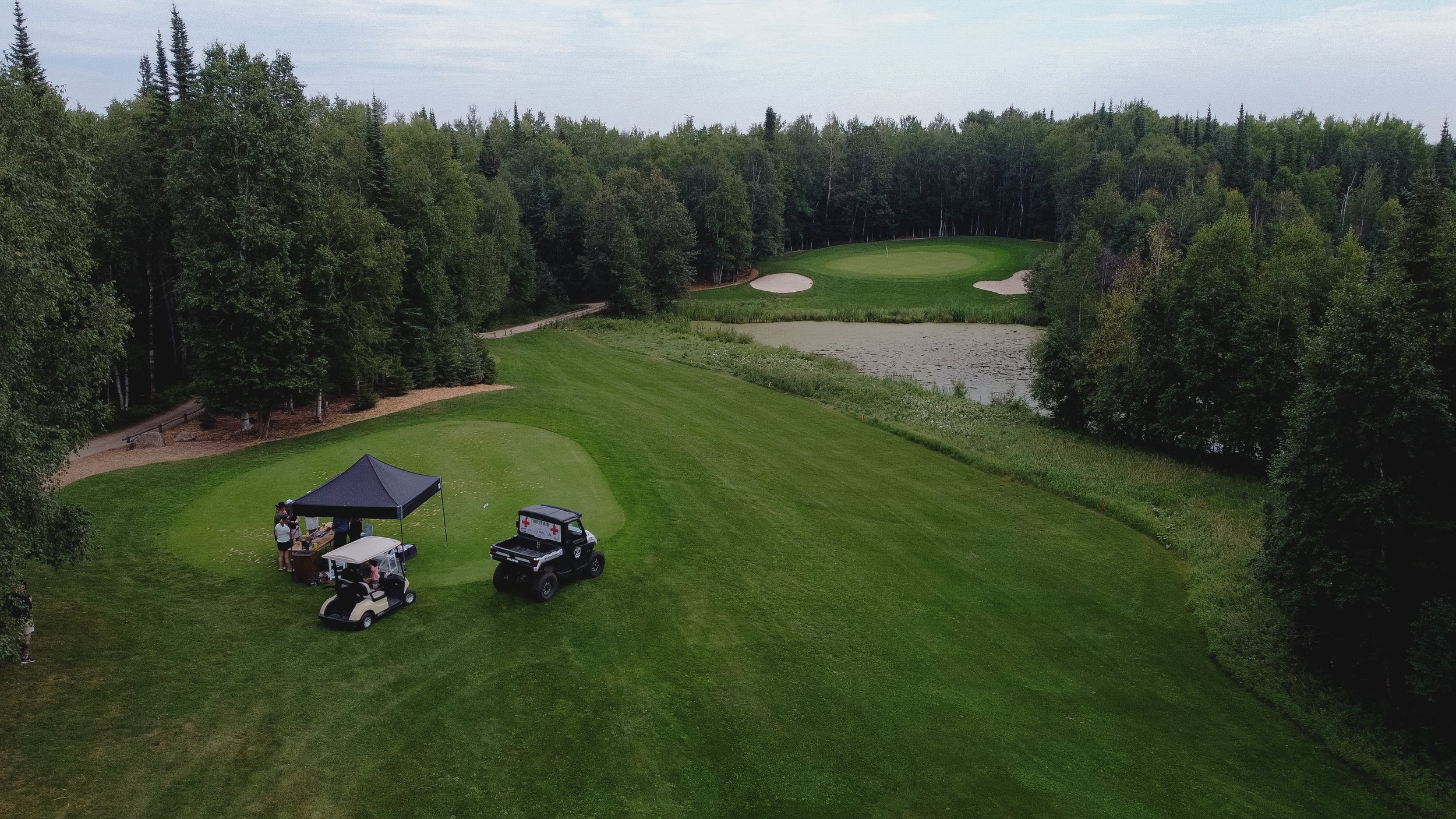 Golf course with a green, sand traps, surrounded by trees, with a pond in the background. There is a tent and vehicles with people gathered near the tent.
