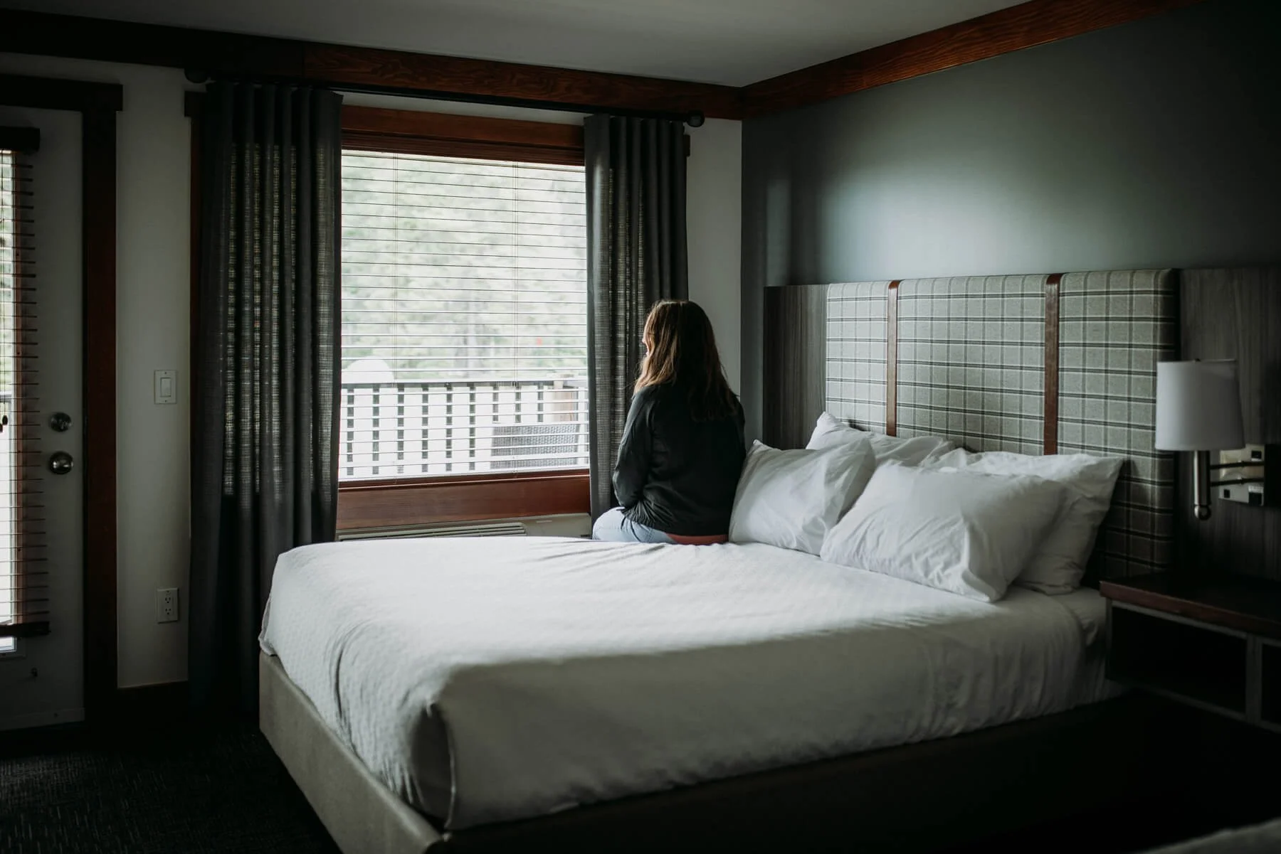 A woman sitting on a bed in a hotel room, looking out the window.