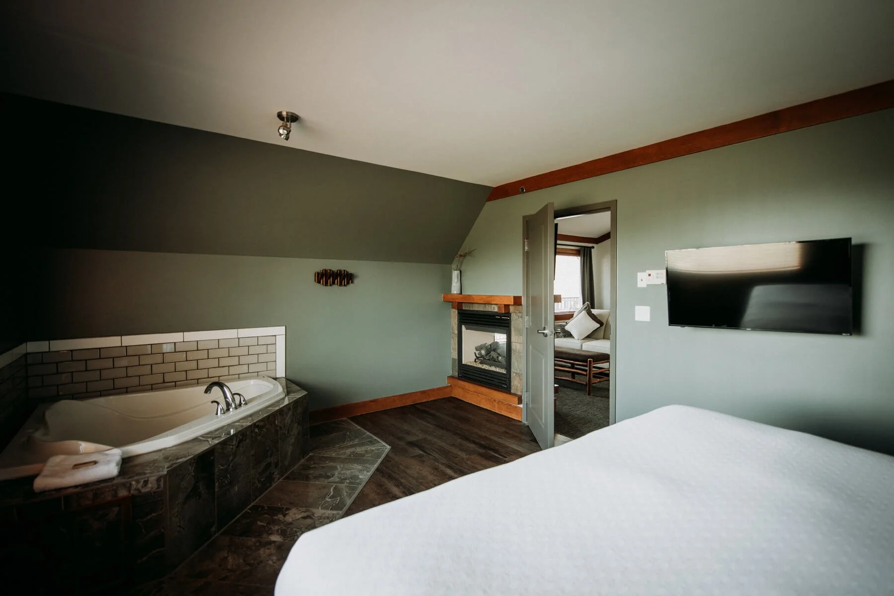 A room with a partial view of a bathtub, a fireplace, a wall-mounted TV, and a bed in the foreground. There's an open door leading to another room with a chair and window.
