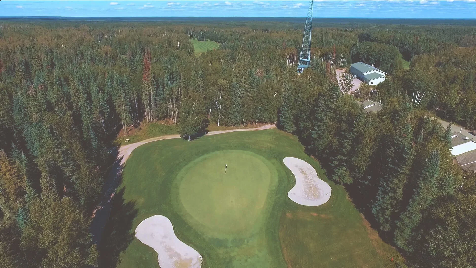 An aerial view of a golf course green surrounded by sand bunkers and trees, with buildings and a tall blue tower in the background.