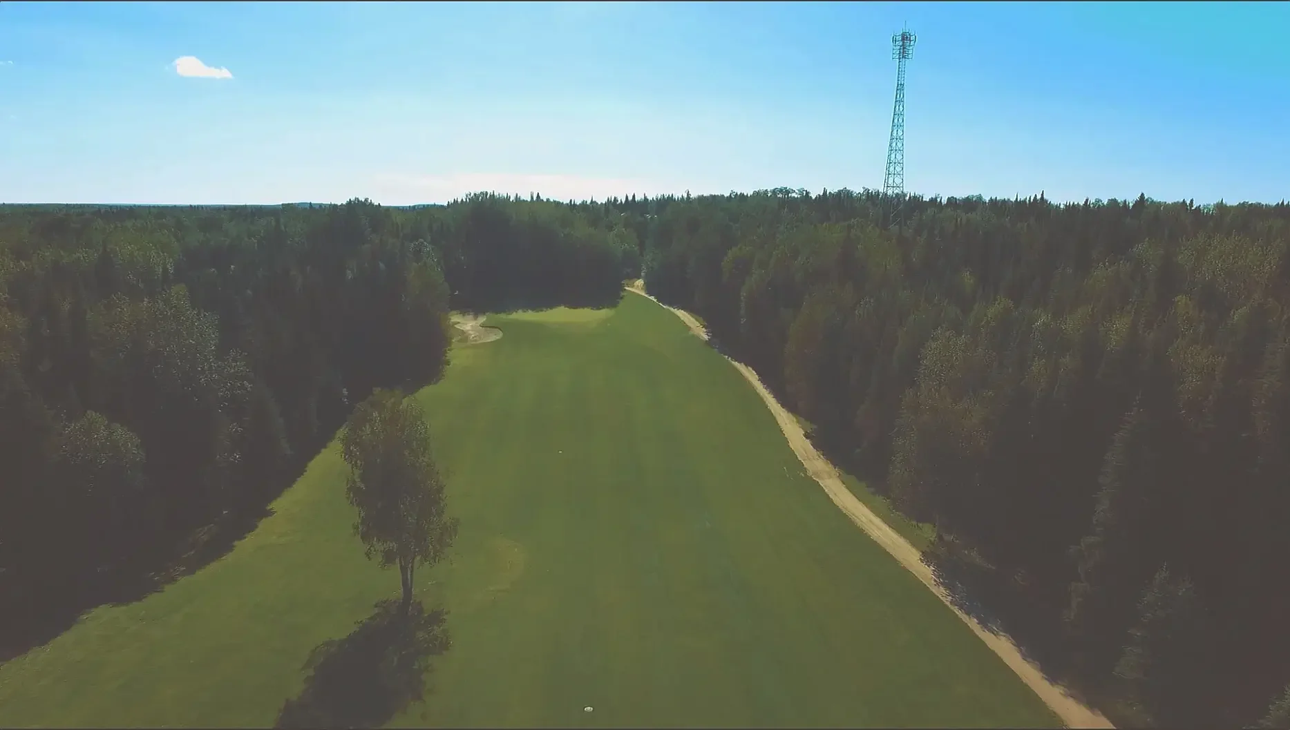 Aerial view of a golf course surrounded by dense forest under a clear blue sky with a small white cloud and a tall communication tower in the distance.