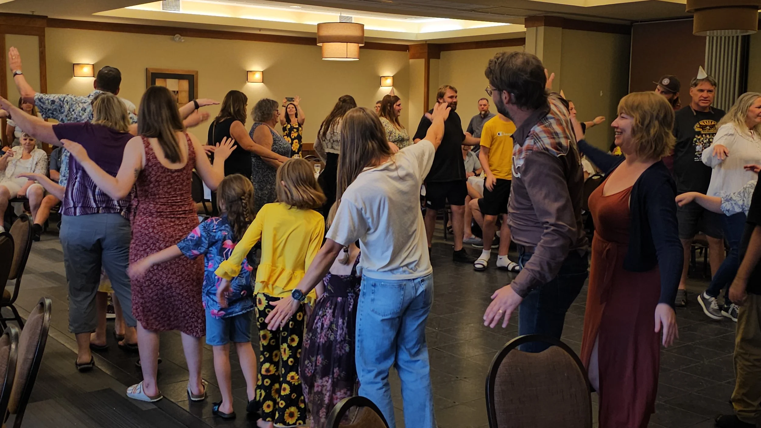 A group of people dancing in a banquet hall with beige walls, wooden accents, and ceiling lights, enjoying a social event.