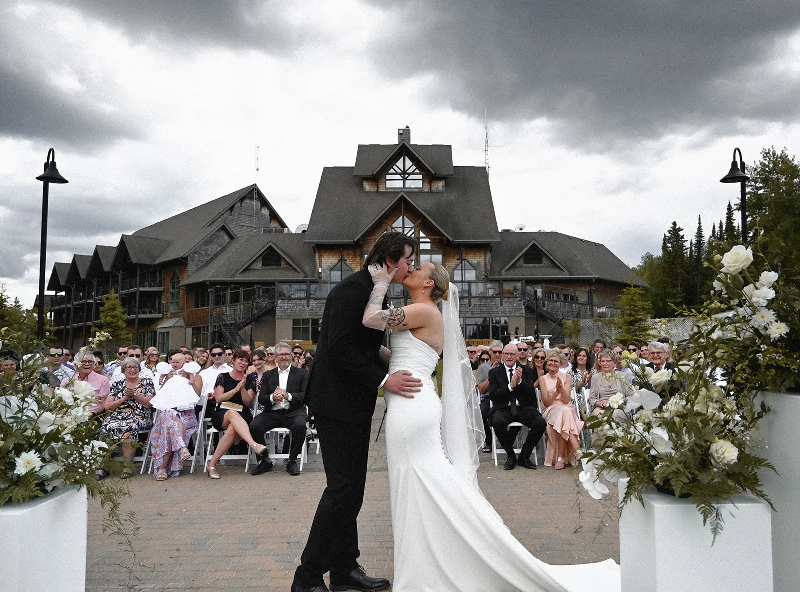 A bride and groom share a kiss during their outdoor wedding ceremony with a large crowd of guests and a rustic building in the background.