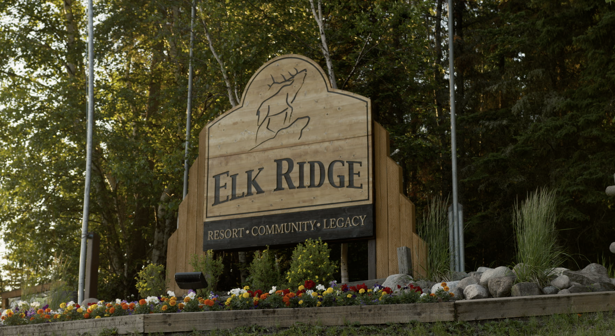 Wooden sign for Elk Ridge Resort, Community, and Legacy, surrounded by colorful flowers in front and trees in the background.