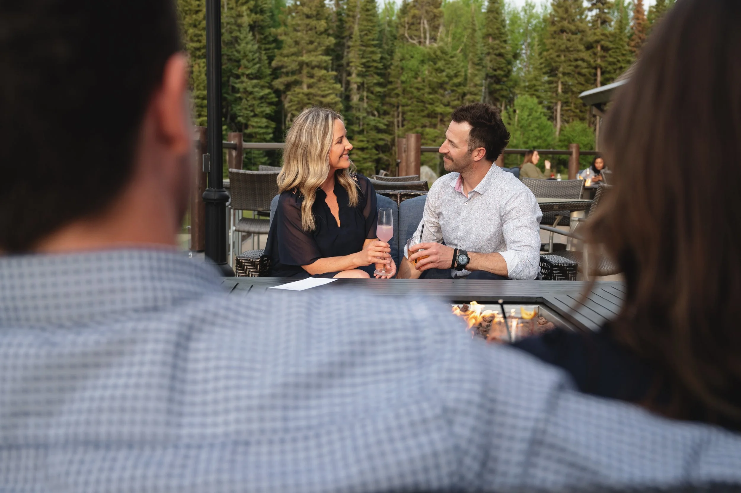 A couple sitting outdoors at a restaurant table, smiling at each other while holding drinks, with a forested background.