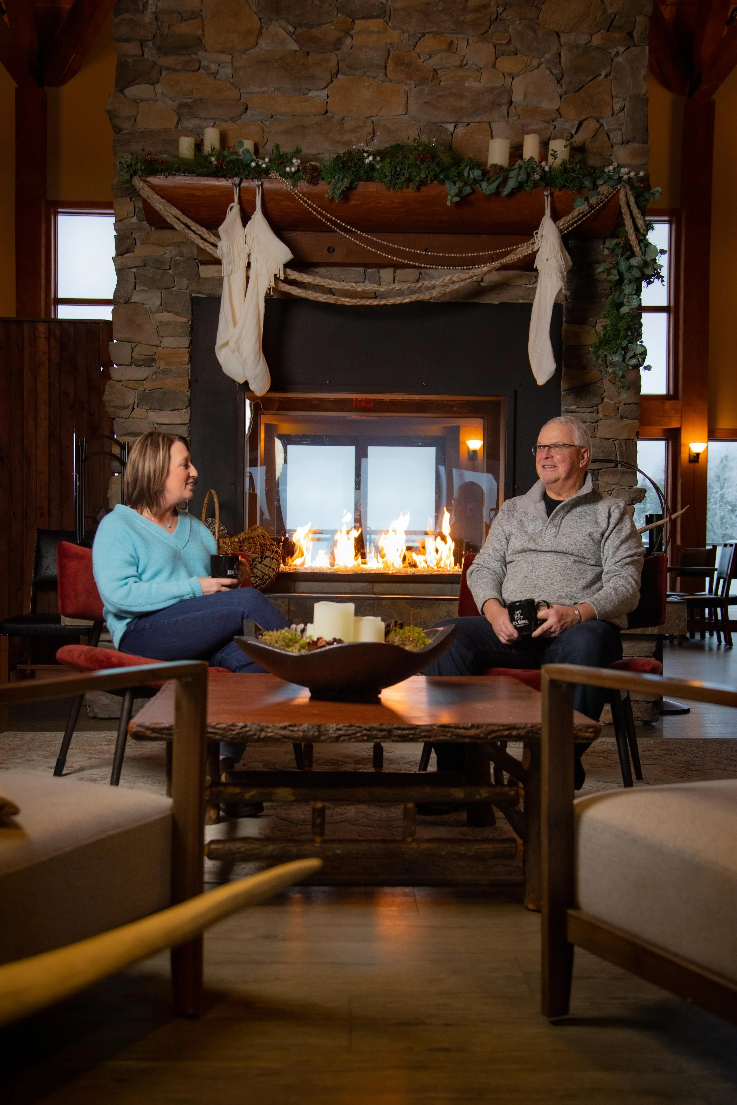 A cozy living room with a stone fireplace decorated for Christmas, featuring stockings hanging from the mantle, candles, greenery, and garlands. Two people are seated near the fireplace, drinking coffee and chatting, with a wooden table in the foreground and a warm, inviting atmosphere.