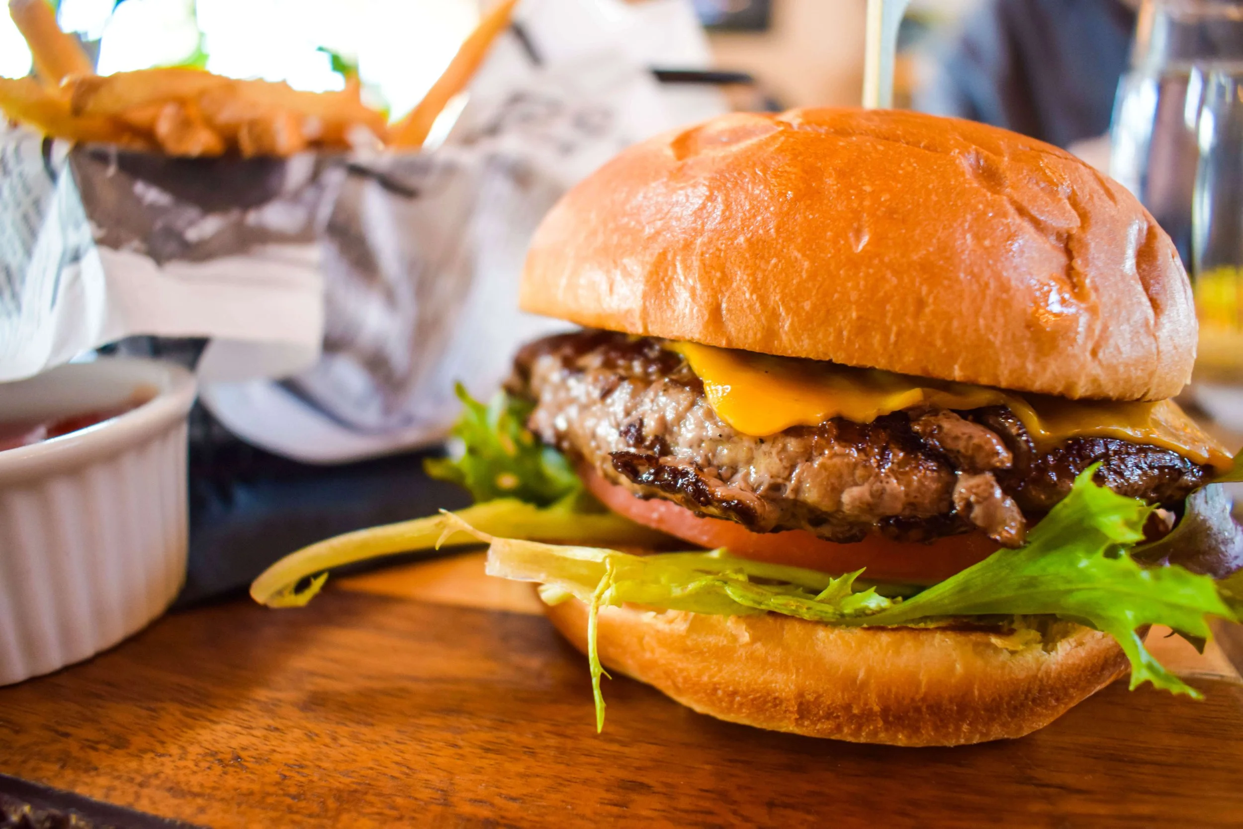 Close-up of a cheeseburger with a beef patty, melted cheese, lettuce and tomato on a toasted bun, with fries and condiment cups behind it.