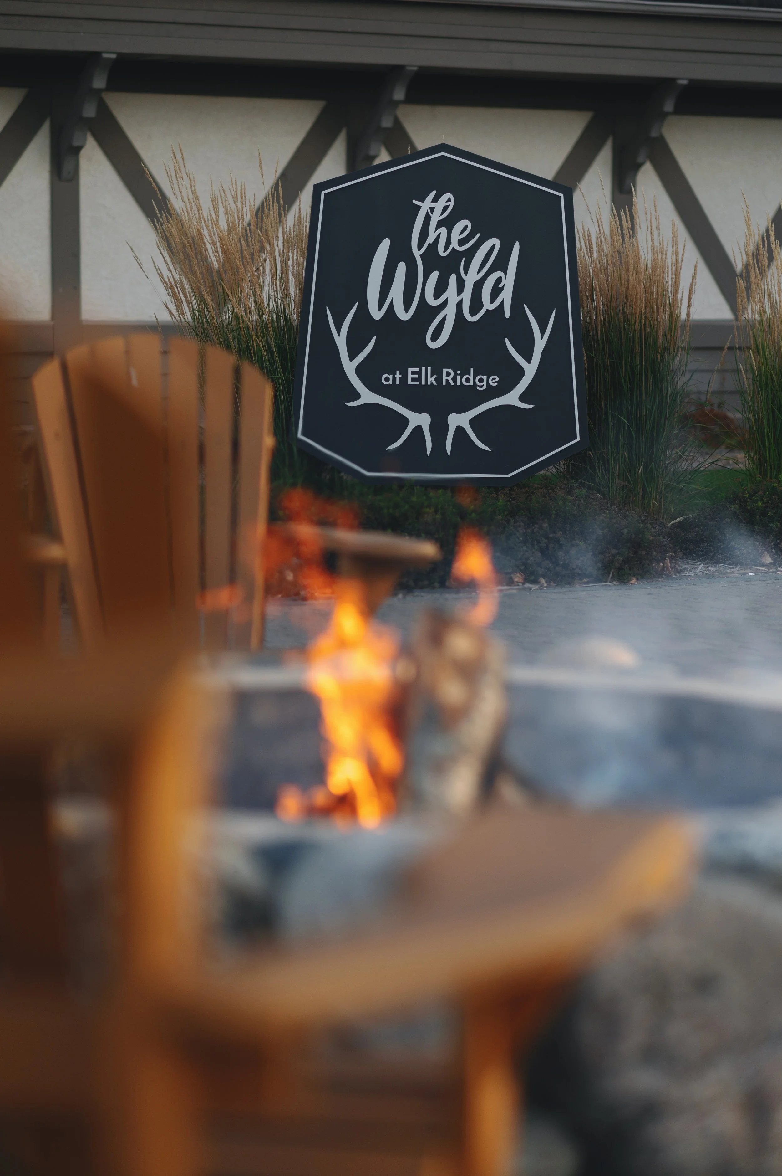 A sign reading &lsquo;the Wyld at Elk Ridge&rsquo; with antler graphics, surrounded by ornamental grasses, with a fire pit burning in the foreground.