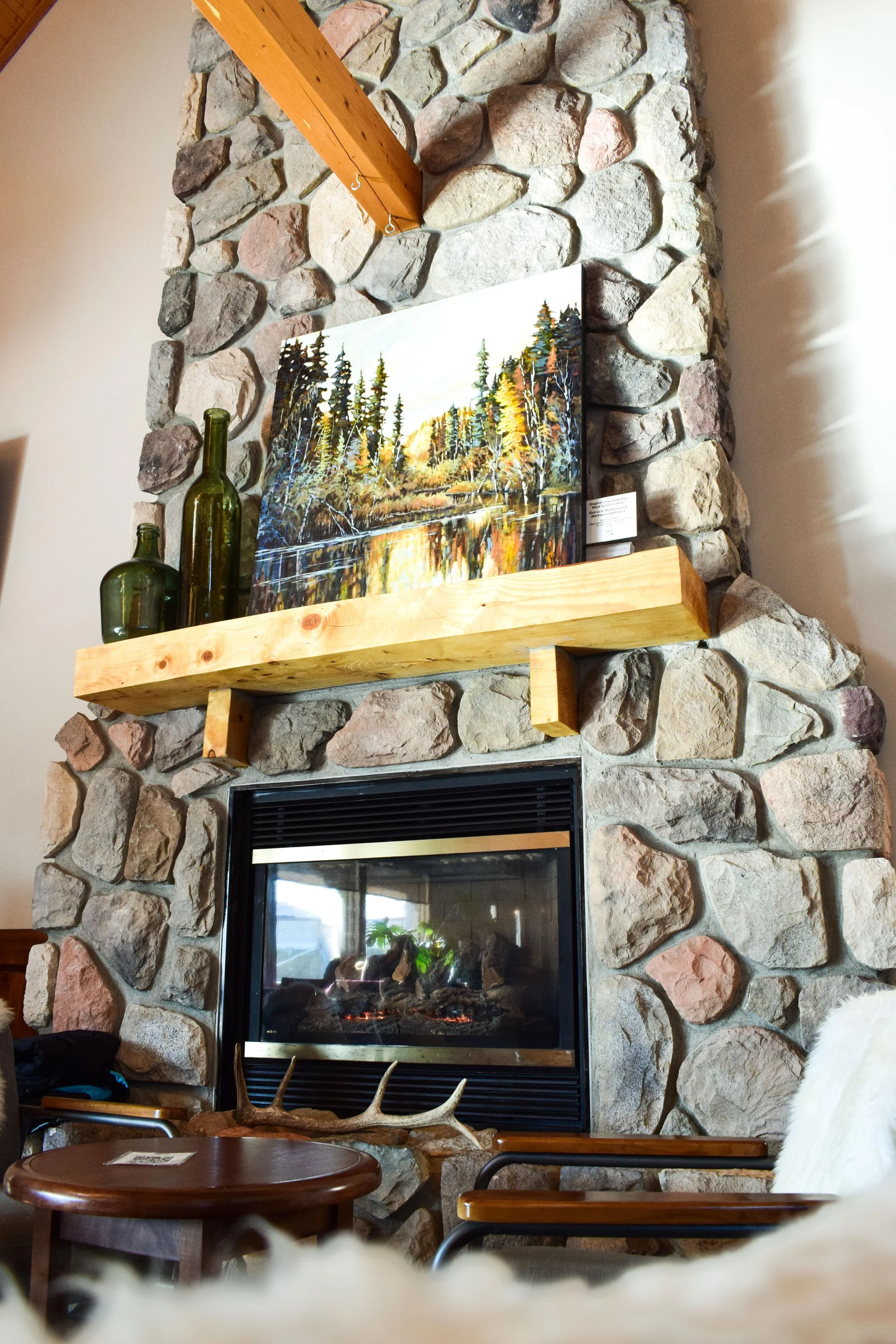 A stone fireplace with two green glass bottles and a painting of a forest and river on the mantle, and a table with an antler on it below.