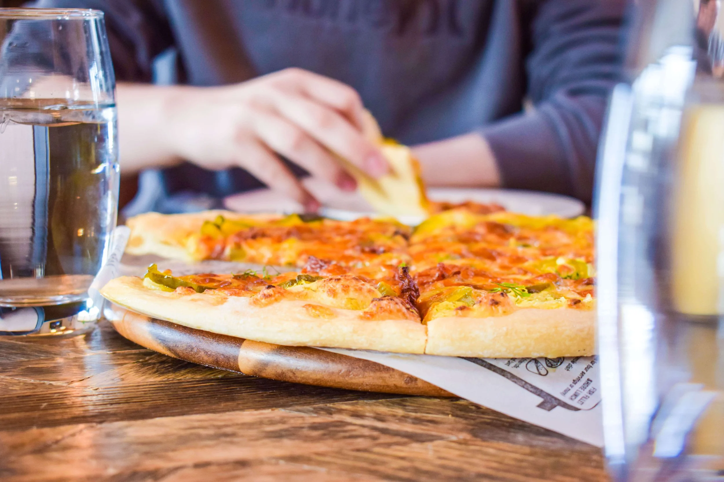 A person at a table lifting a slice from a fresh pizza topped with green peppers and cheese.