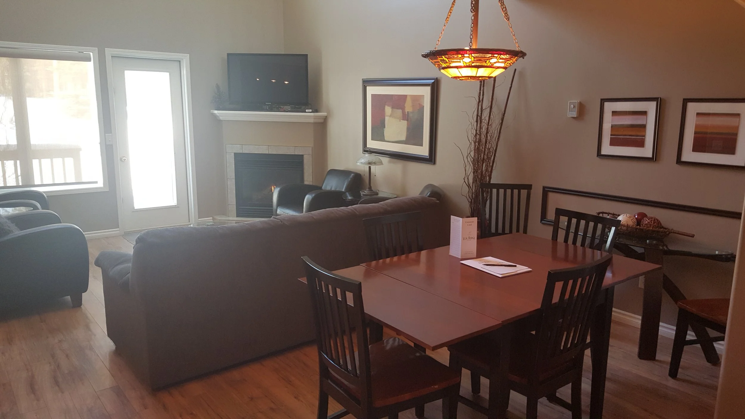 Living room with gray couches, a black armchair, a fireplace, wall art, a wooden dining table with chairs, a hanging light, and a basket of decorative balls.