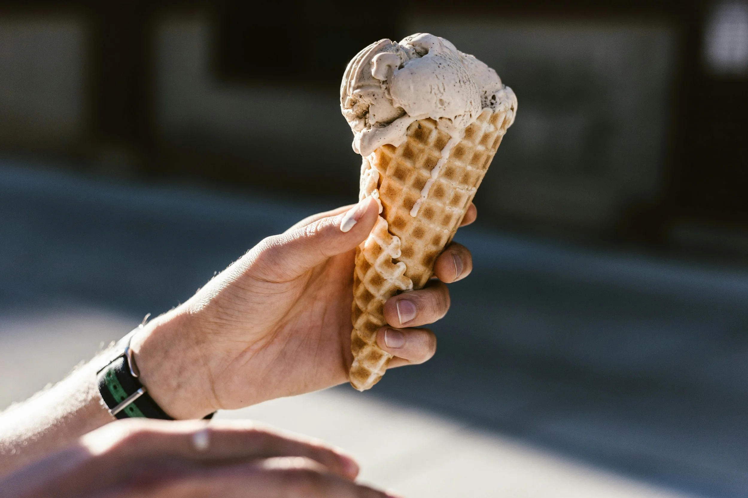 A hand holding a waffle cone with two scoops of ice cream, with a blurred background.