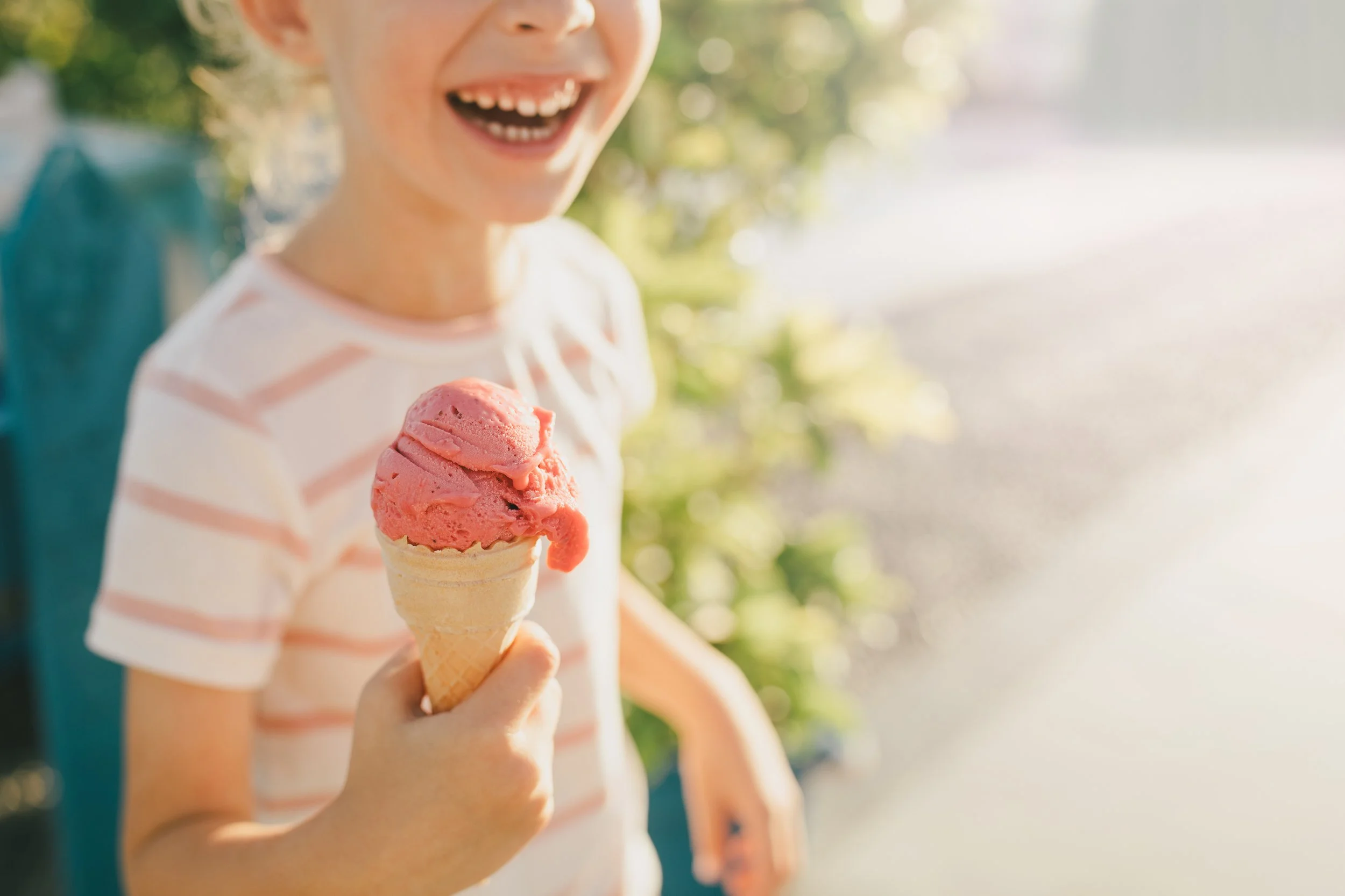 A child smiling and holding a cone of pink ice cream outdoors, wearing a white and pink striped shirt.