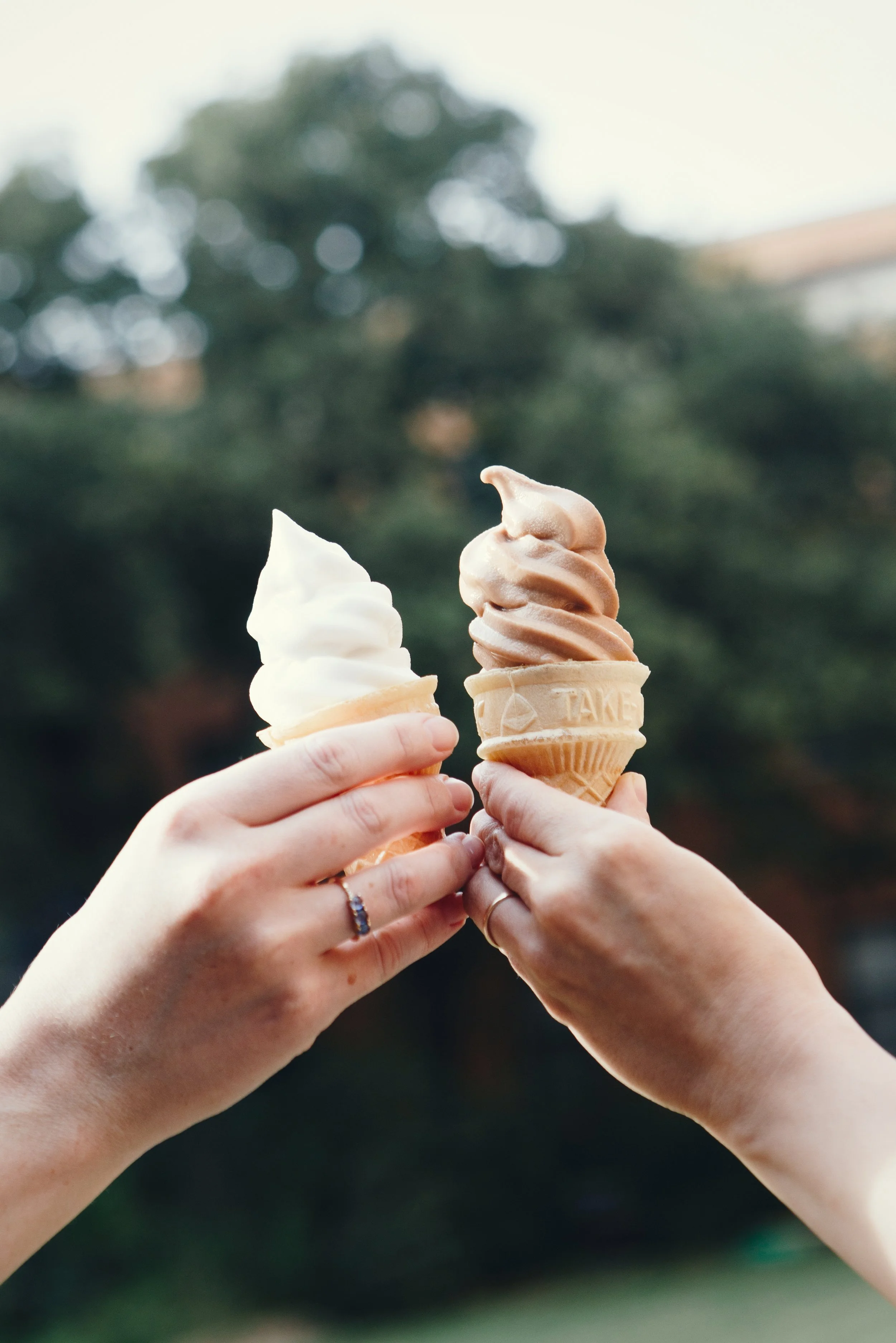 Two hands holding ice cream cones, one vanilla and one caramel, outdoors against a blurred background of trees.