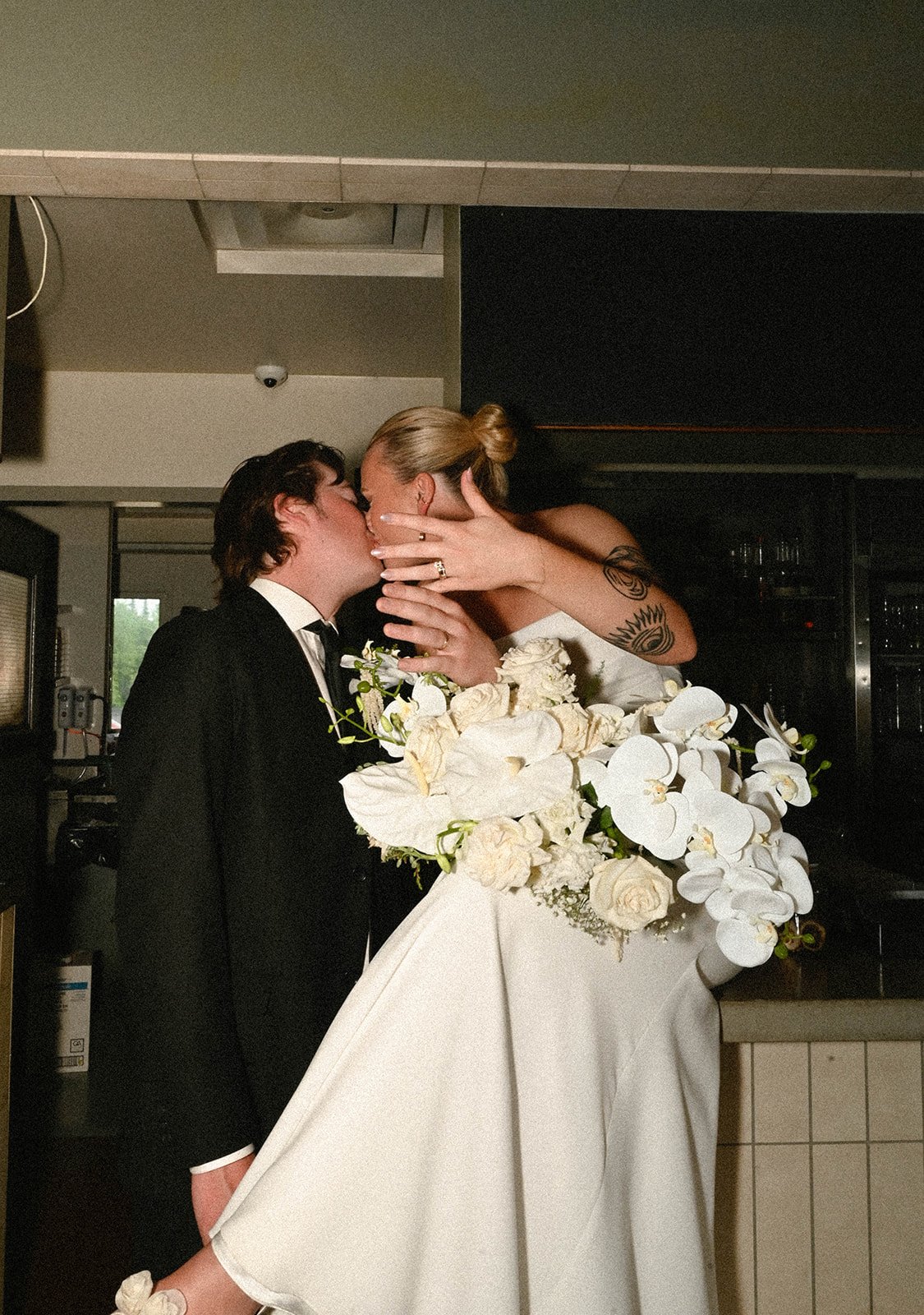 A bride and groom share a kiss at their wedding reception, with the bride's arms around the groom and the groom holding a large bouquet of white flowers.