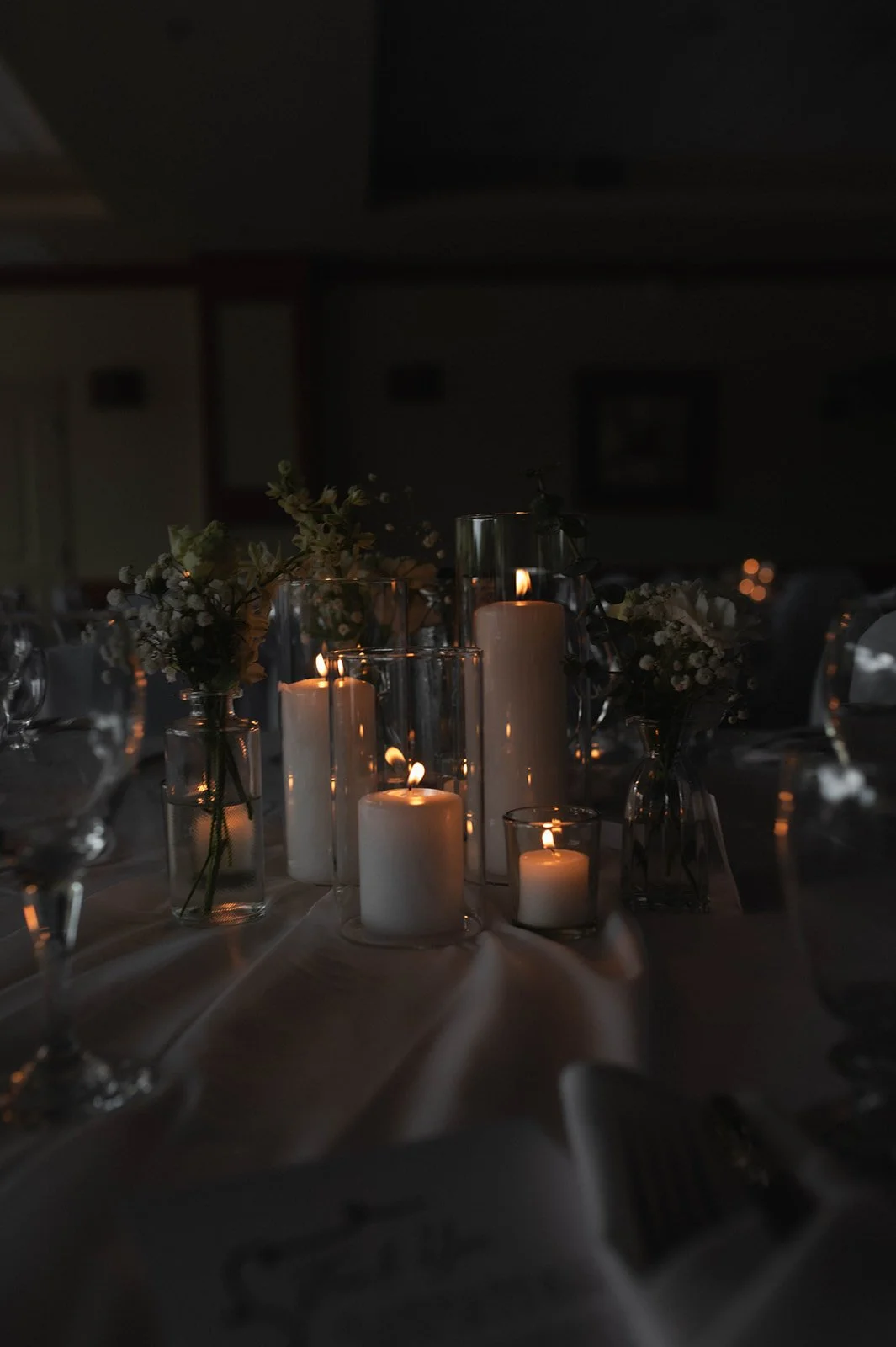 A table decorated with lit candles in glass holders and small floral arrangements in vases during a dimly lit setting, likely for a special event or dinner.
