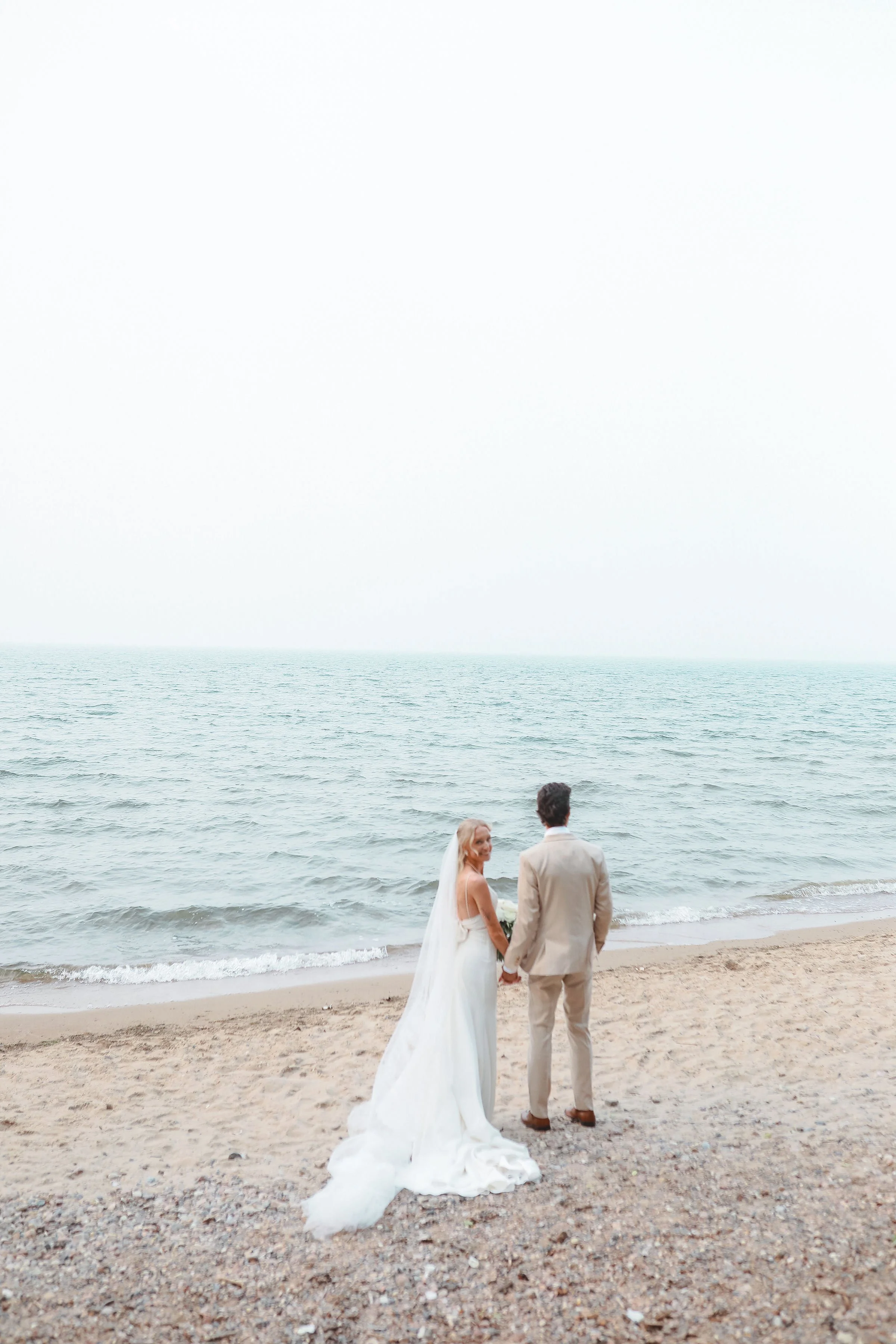 A bride and groom holding hands on a beach, facing the ocean with waves, sandy shore, and an overcast sky.
