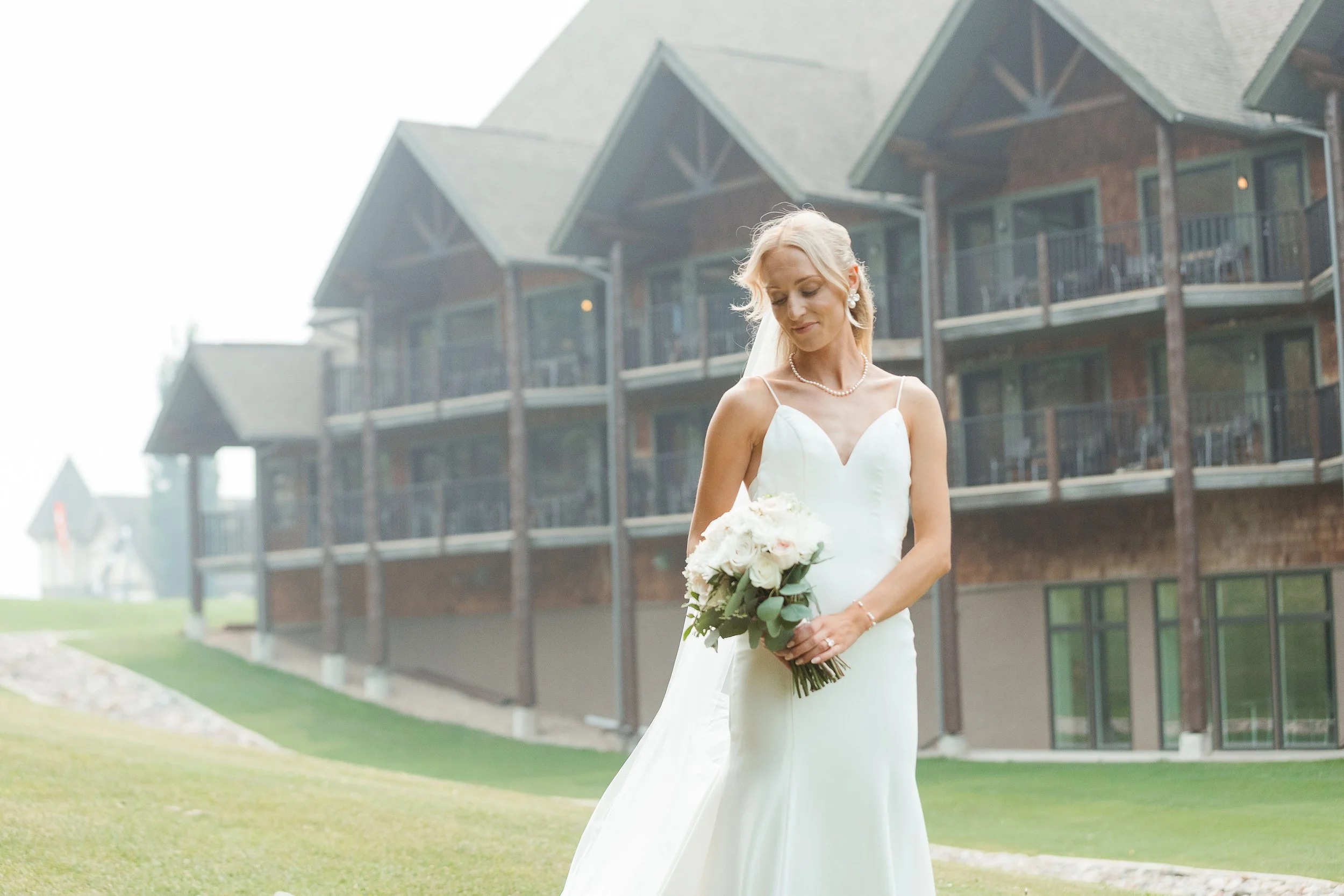 A bride in a white wedding dress holding a bouquet of flowers, standing outdoors in front of a modern townhouse-style building with balconies.