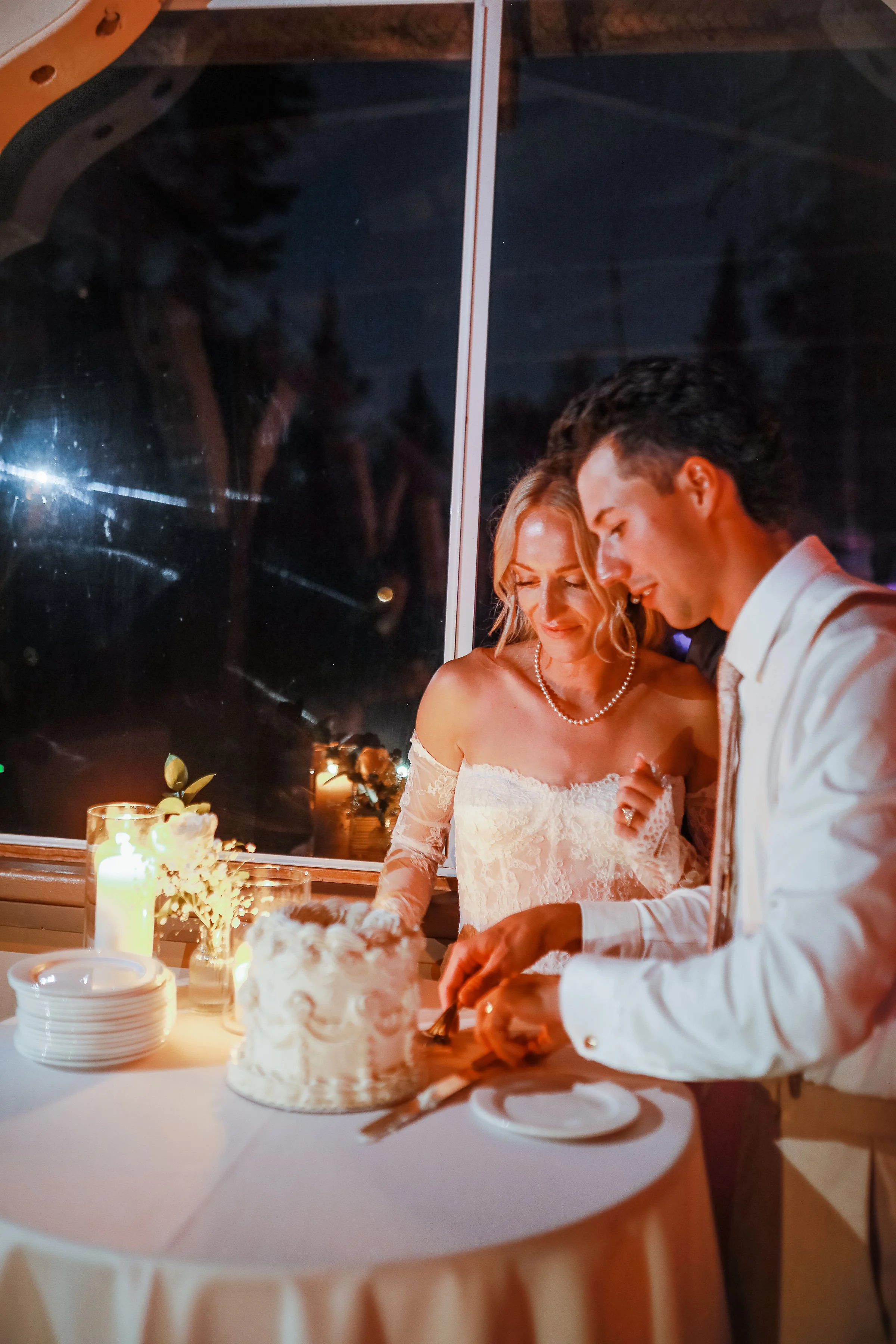 A bride and groom cutting a wedding cake during their wedding reception at night, with candles and floral decorations on the table.