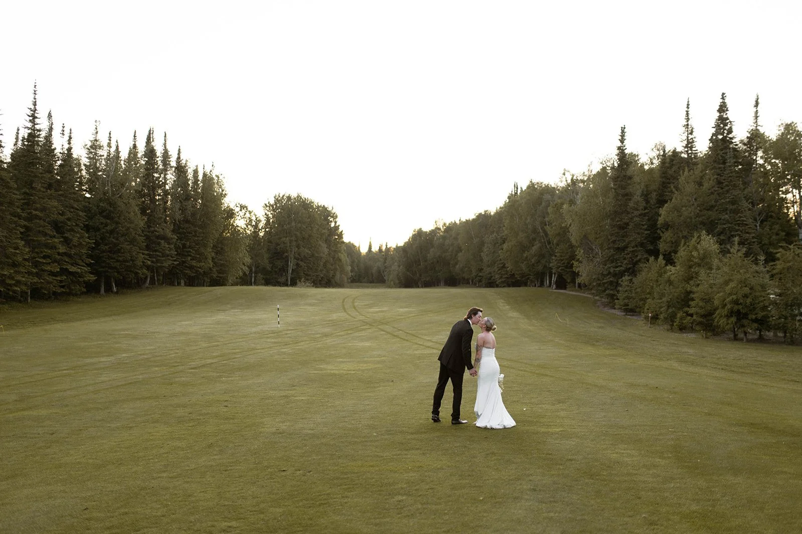 A bride and groom holding hands and kissing on a golf course with trees in the background.