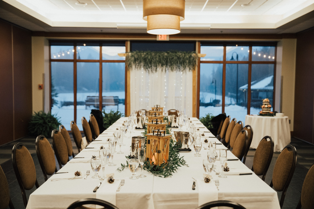 Decorated indoor wedding or event room with a long table set with glassware, lanterns and greenery.