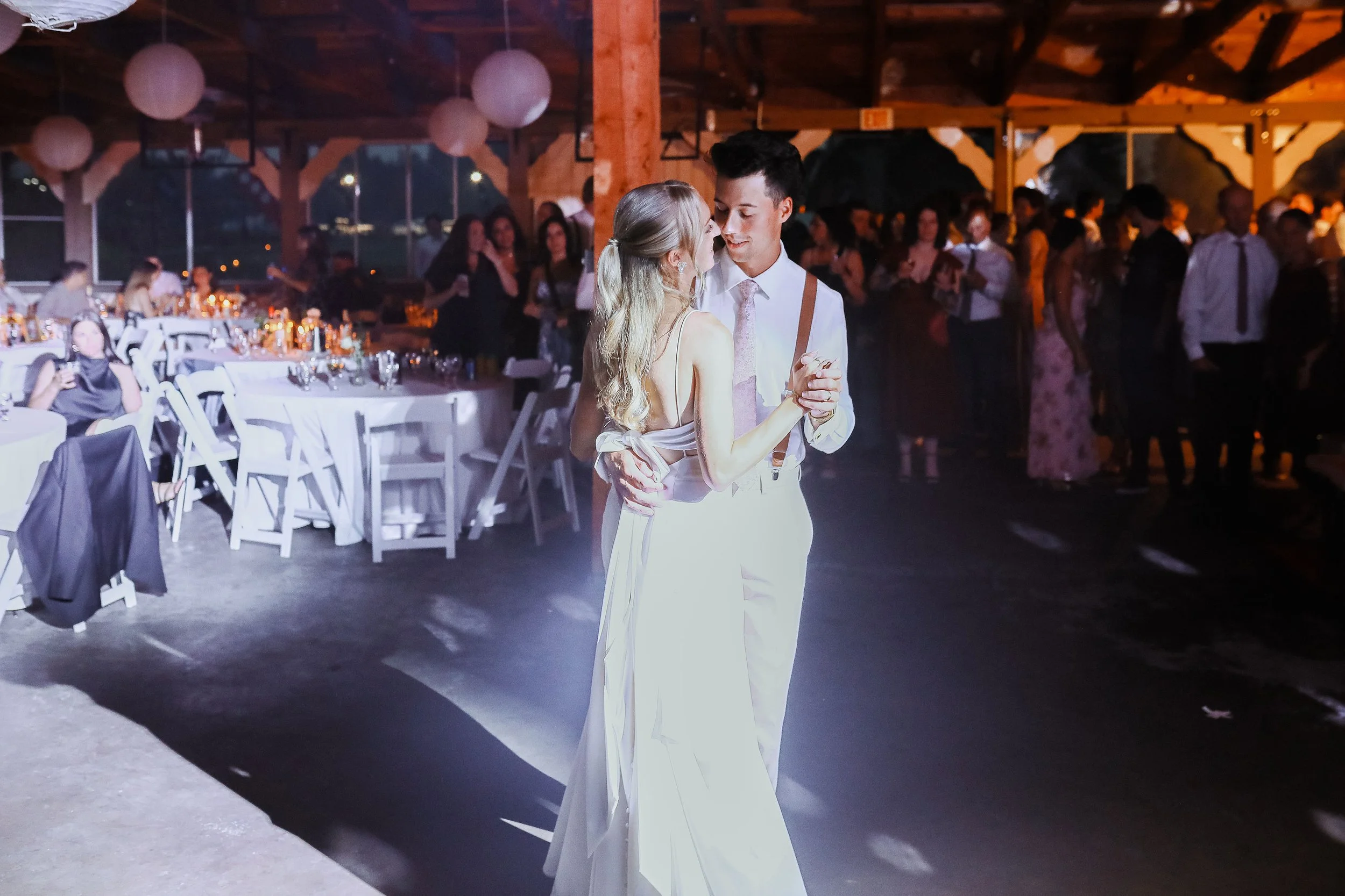 A couple dancing at a wedding reception, with guests in the background, decorated with string lights.