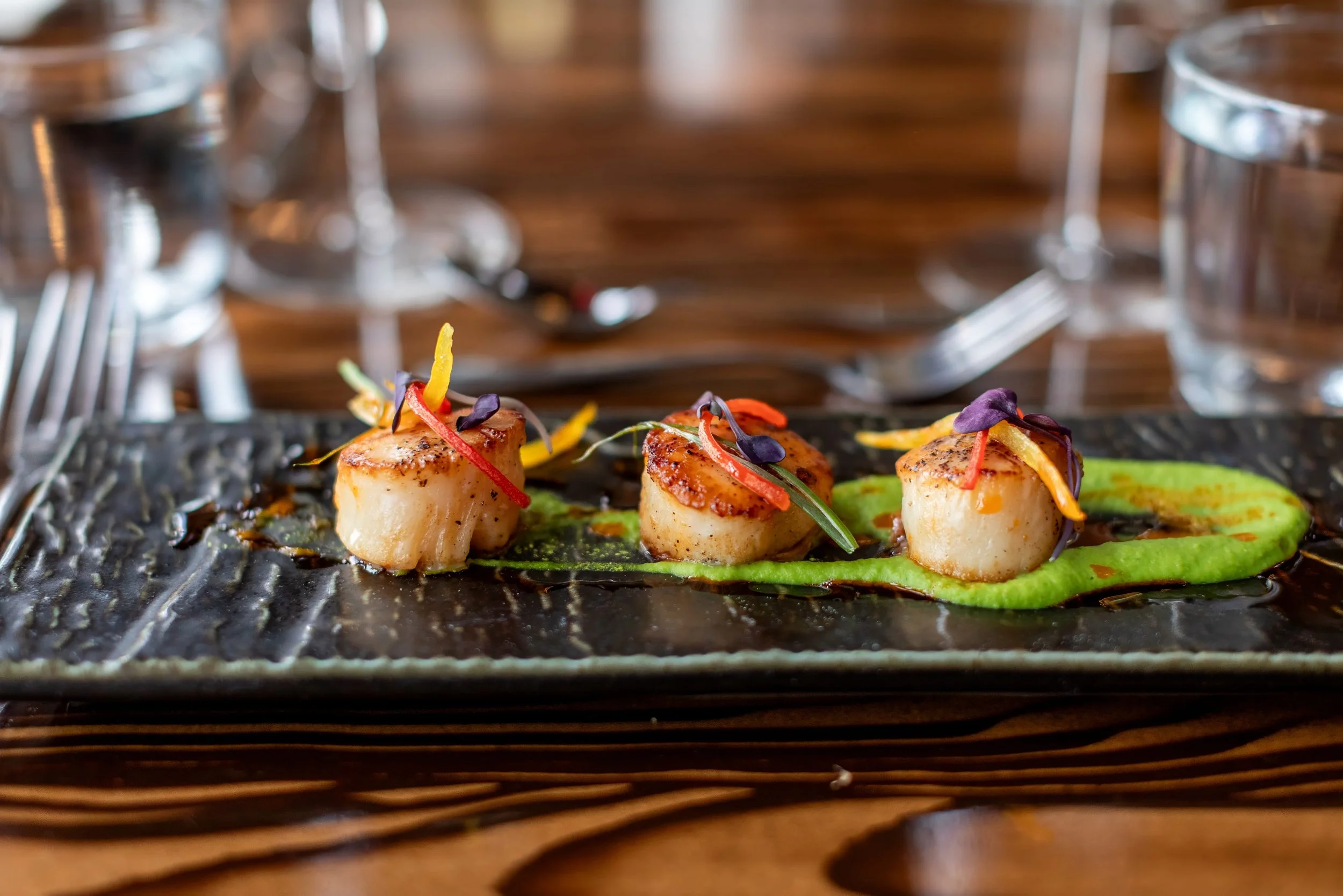Three seared scallops garnished with microgreens and thin strips of colorful vegetables on a slate plate.