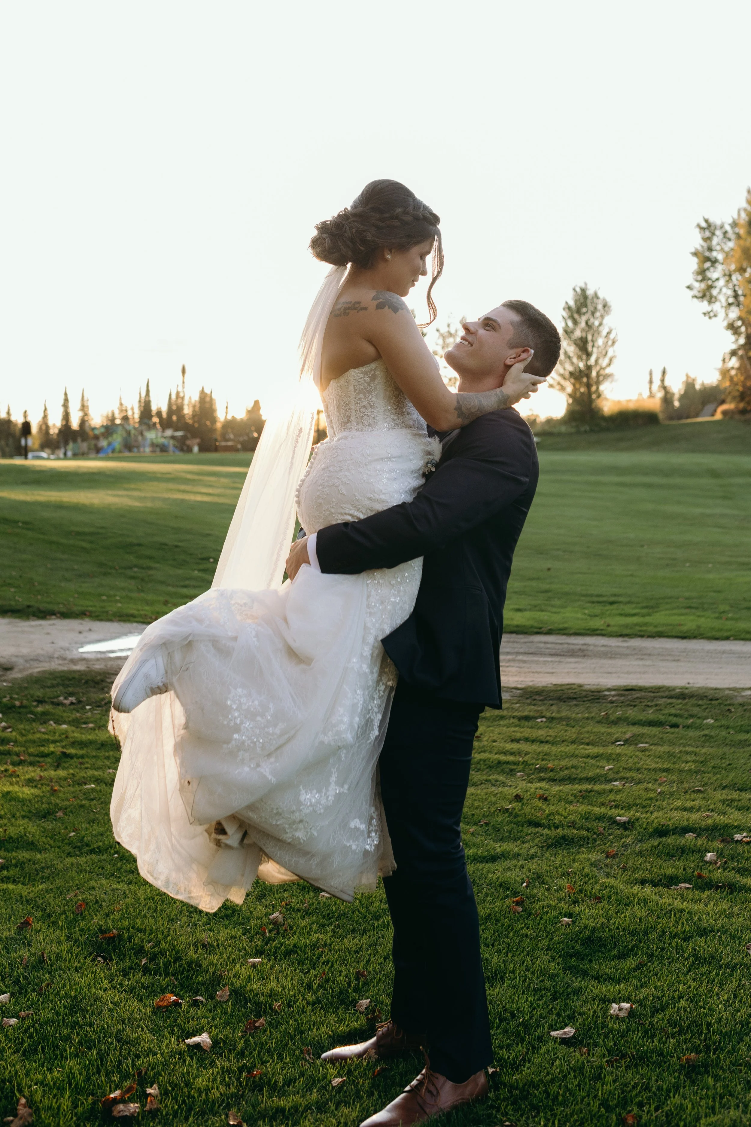 A groom in a black suit and brown shoes lifts a bride in a white lace wedding dress with a long train.